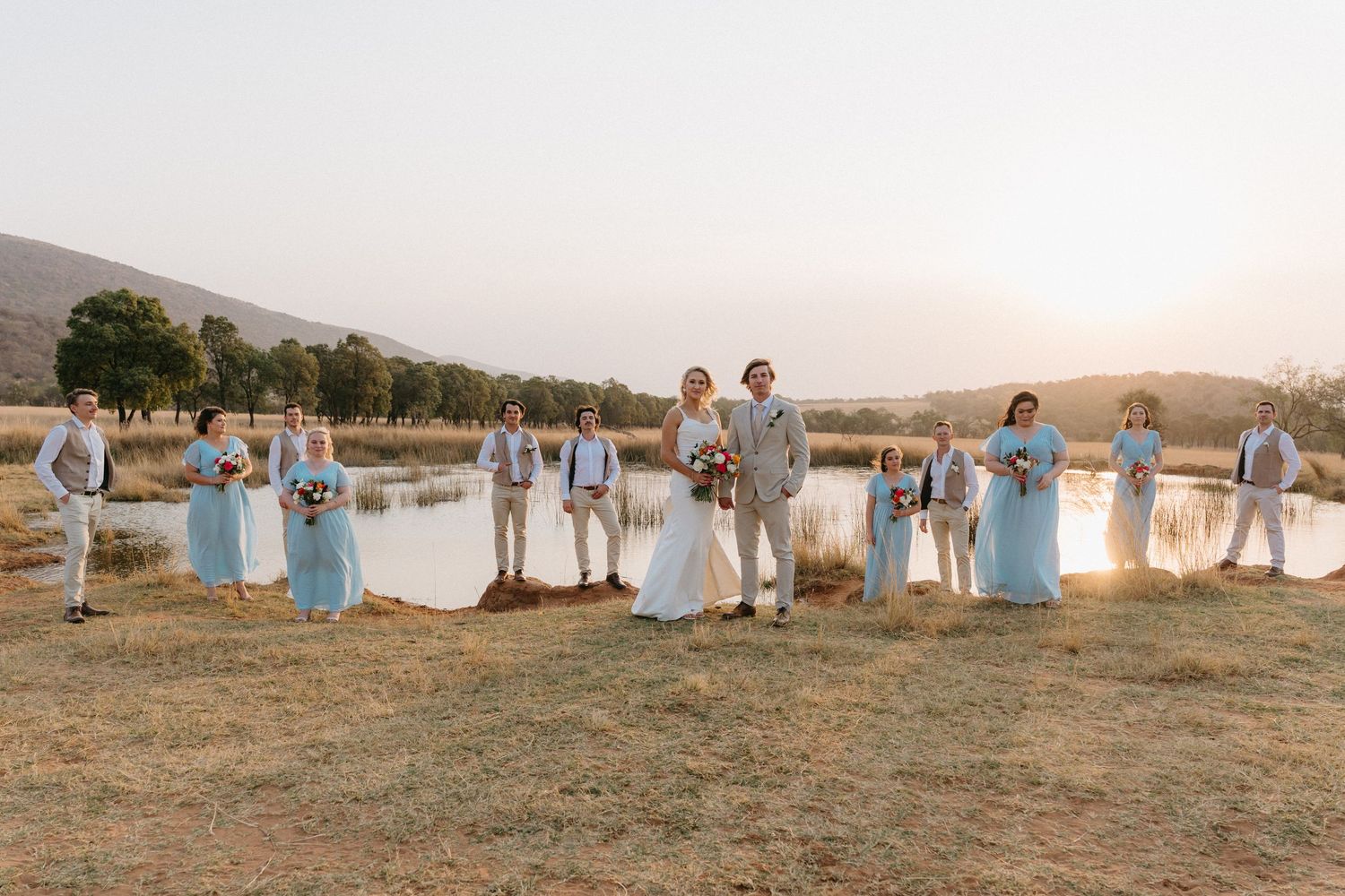 Wedding party in light blue dresses and beige suits pose at sunset by a lake.