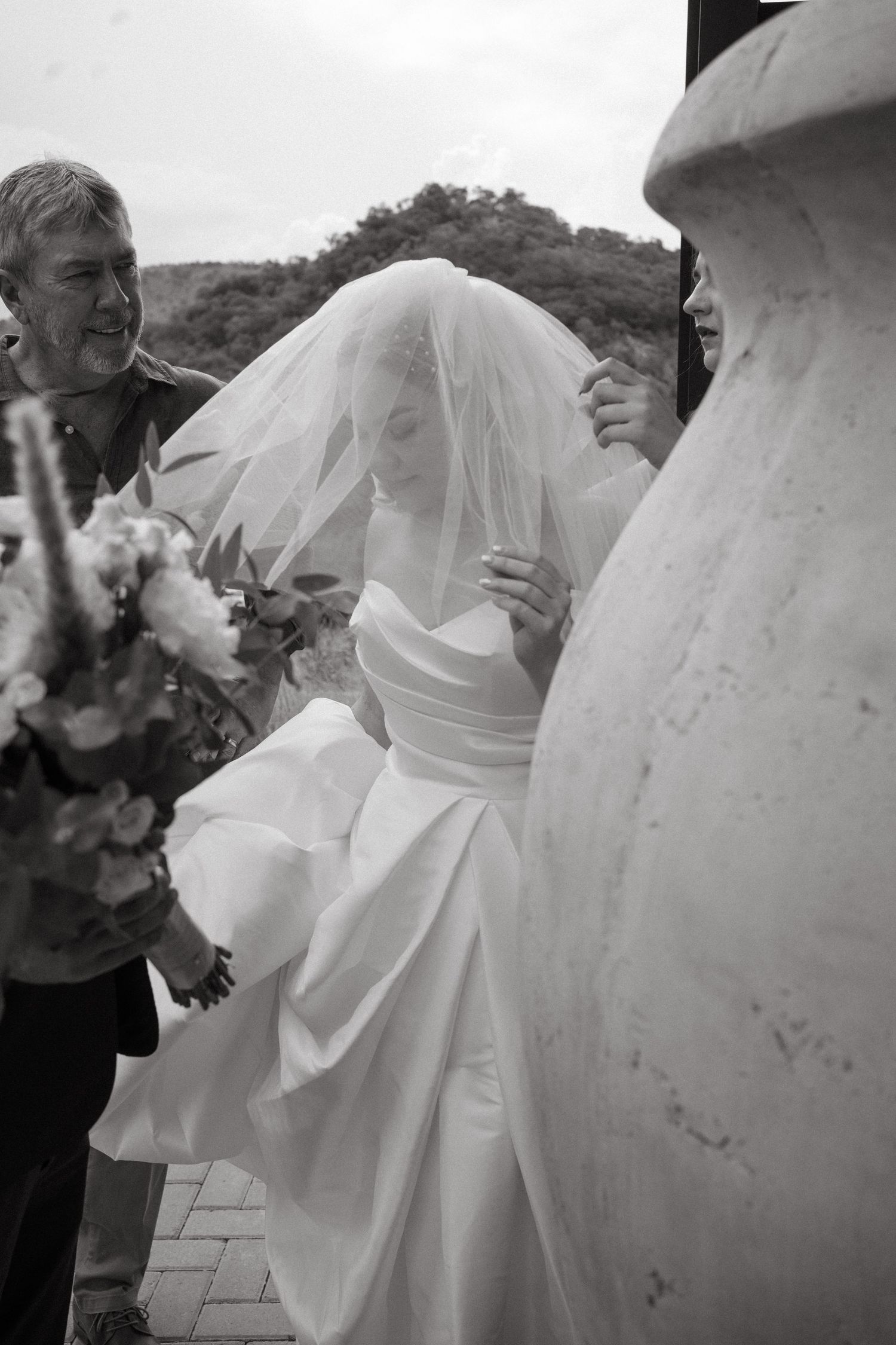 Black and white photo showing a wedding party with flowing white bridal gown and bouquet outdoors.