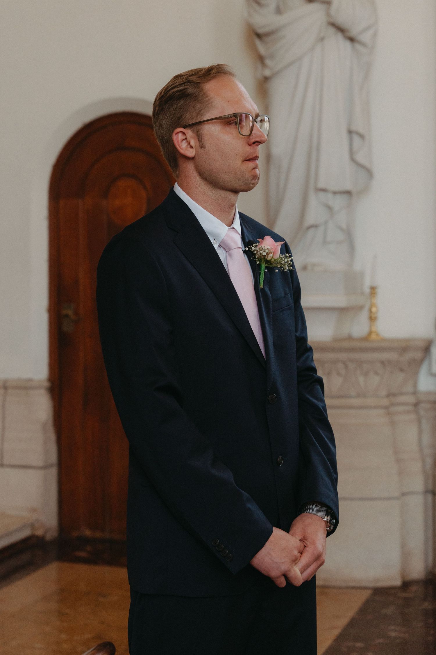 A groom in a navy suit and glasses stands solemnly in a church interior with a pink boutonniere.