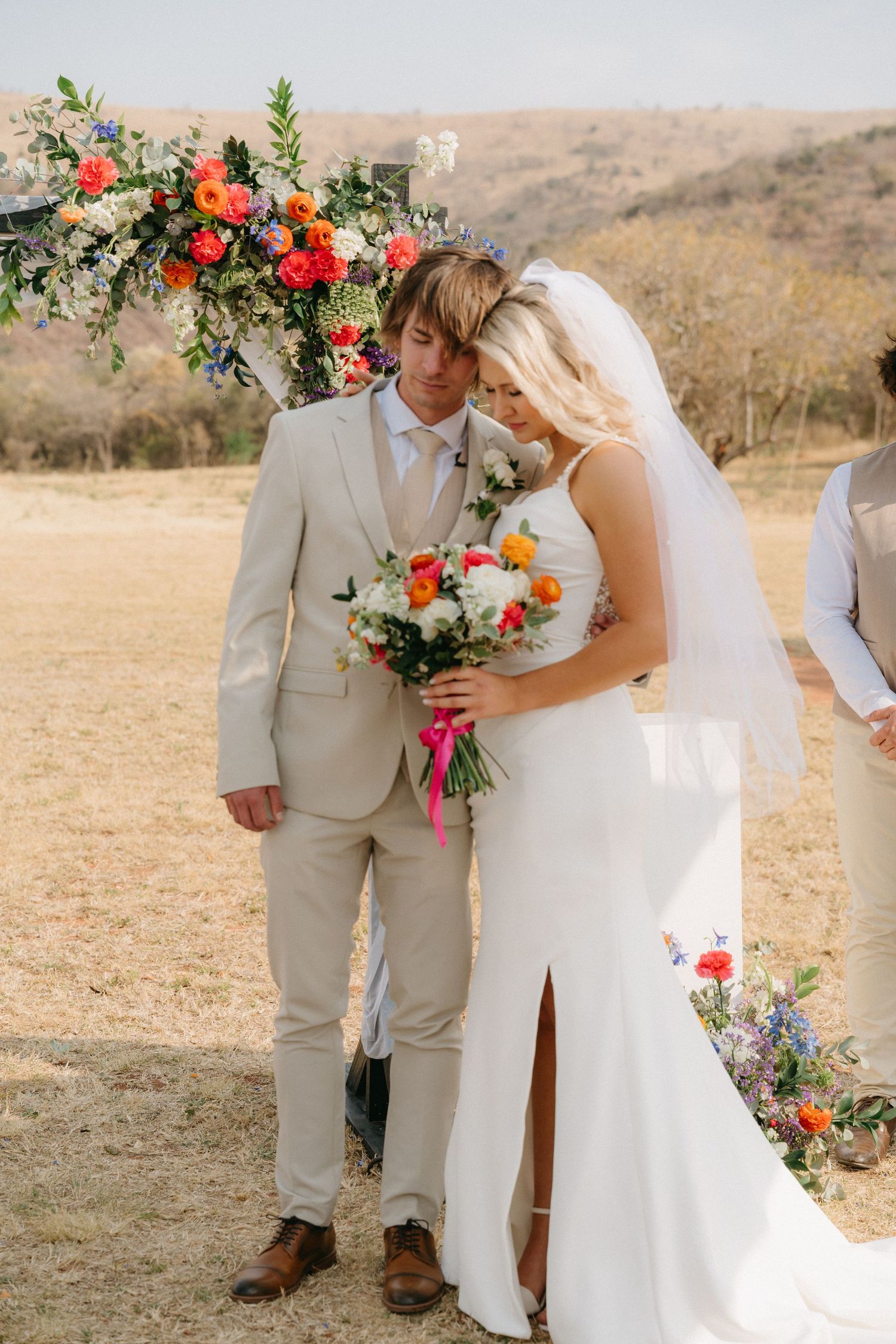 Wedding couple in tan suit and white dress pose with vibrant coral and orange floral arrangements outdoors.