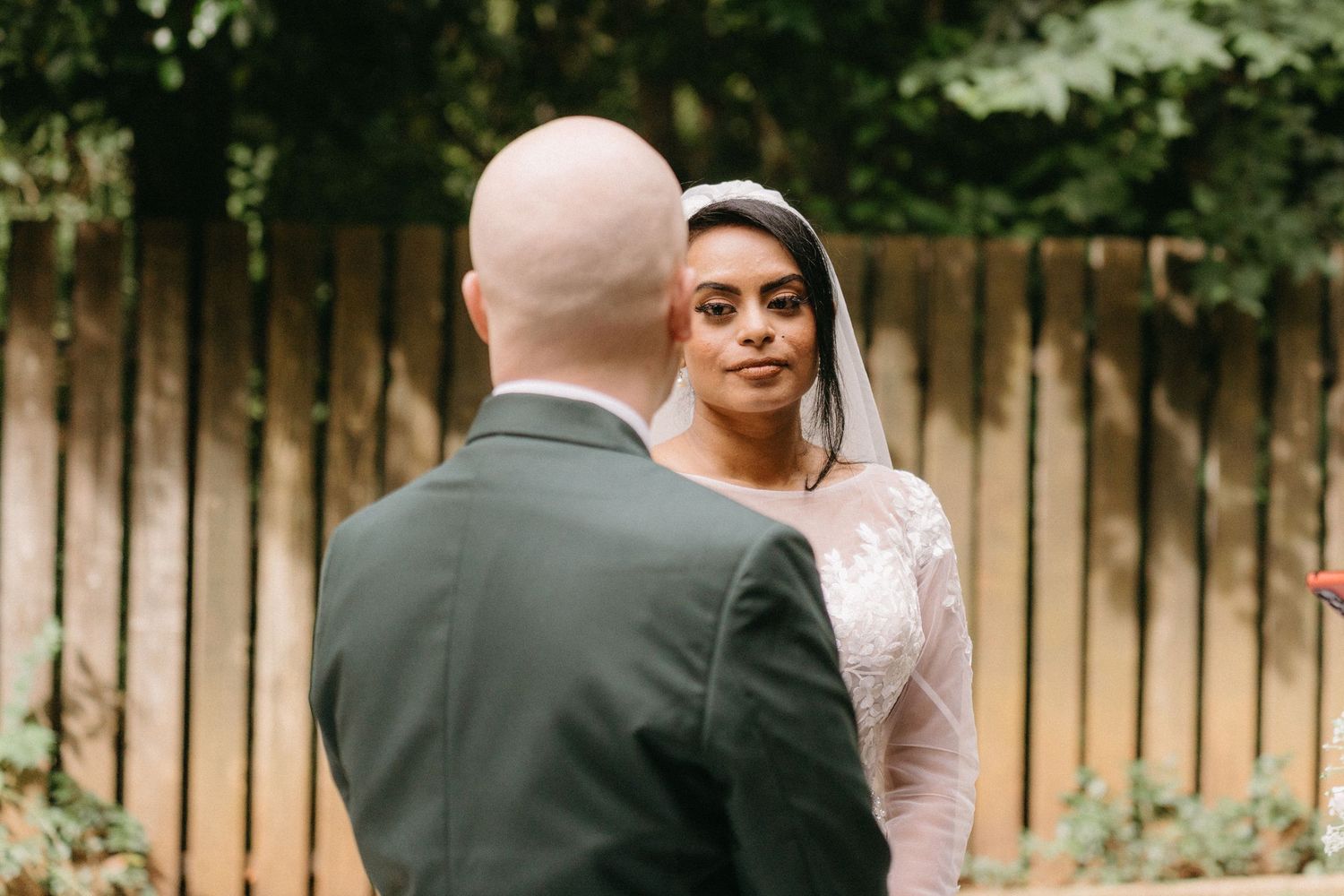 A couple shares a beautiful outdoor wedding moment in front of a wooden fence in an intimate garden setting.