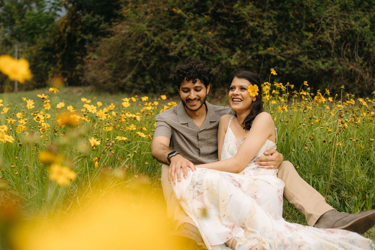 Couple laughing together while sitting in a meadow with blooming yellow flowers.