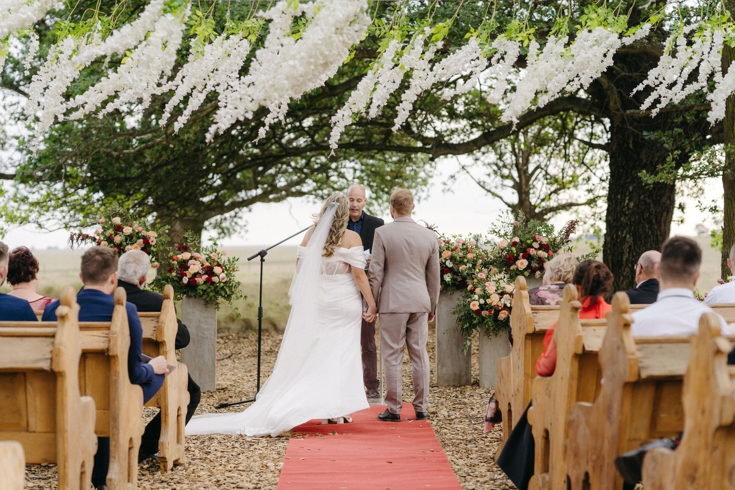 Romantic outdoor wedding ceremony under a floral-draped pergola with wooden pew seating and red aisle runner.