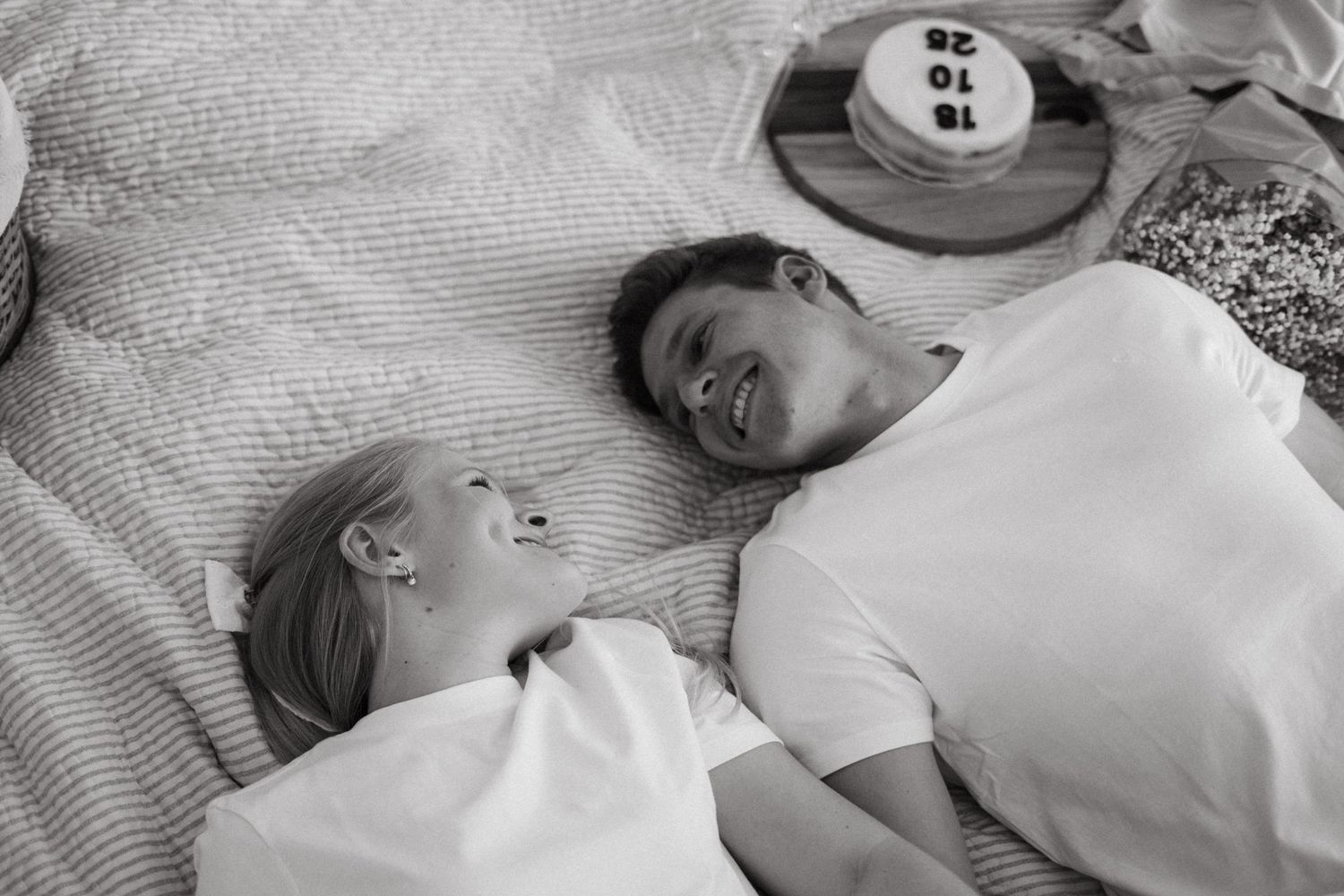Two people laying down on a white bedding surface laughing joyfully together in black and white photography.