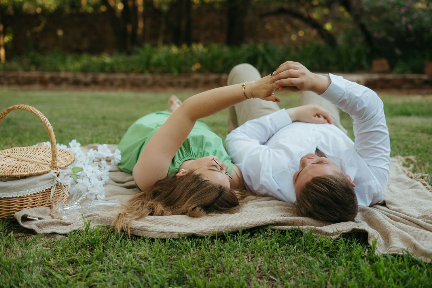 A couple lying on a blanket in a garden with picnic basket and flowers nearby.