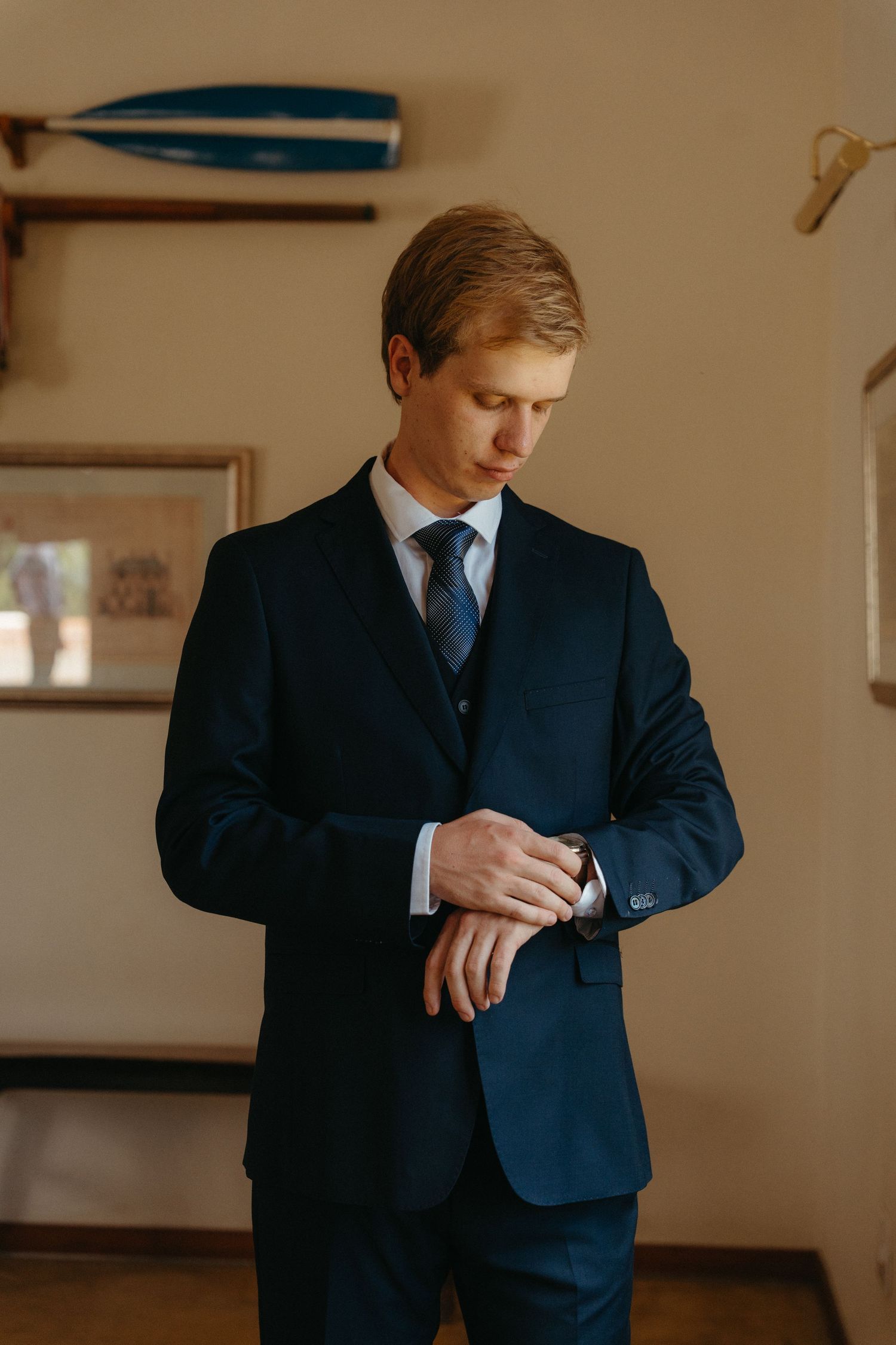 A person in a navy blue suit adjusts their sleeve cuff while standing in a warmly lit interior space.