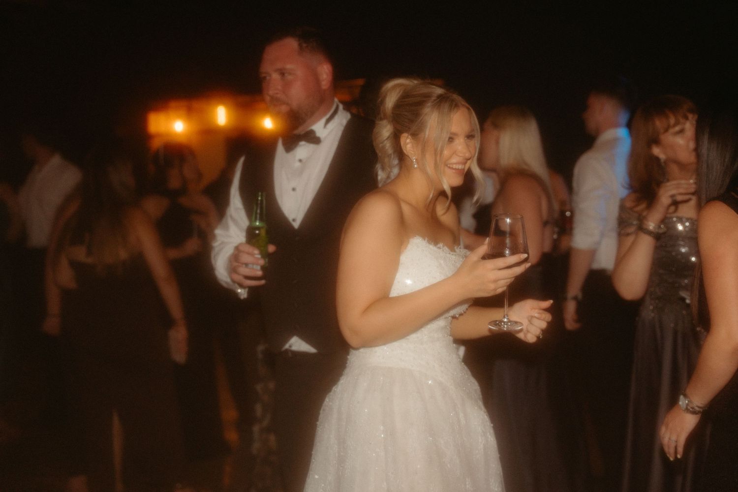 People dancing and celebrating at a wedding reception in a dimly lit venue with warm lighting.