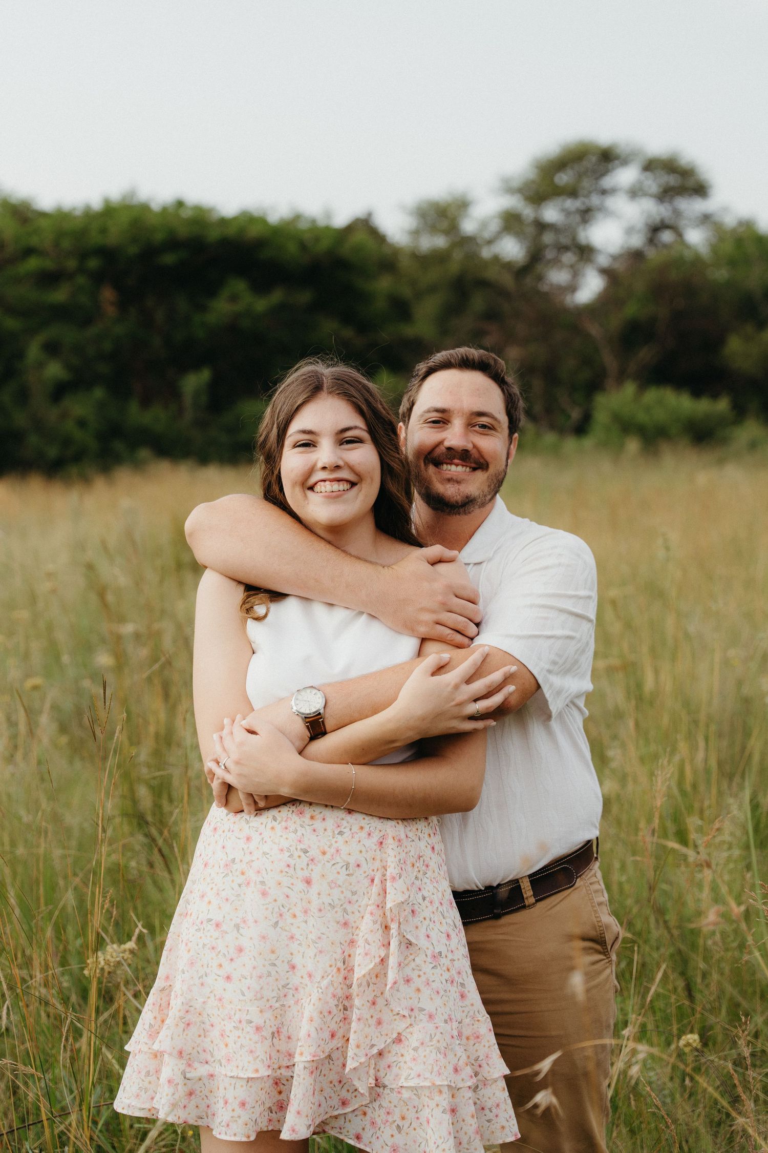 Happy couple embracing in a sunny meadow, woman in floral skirt and man in white shirt smiling outdoors.