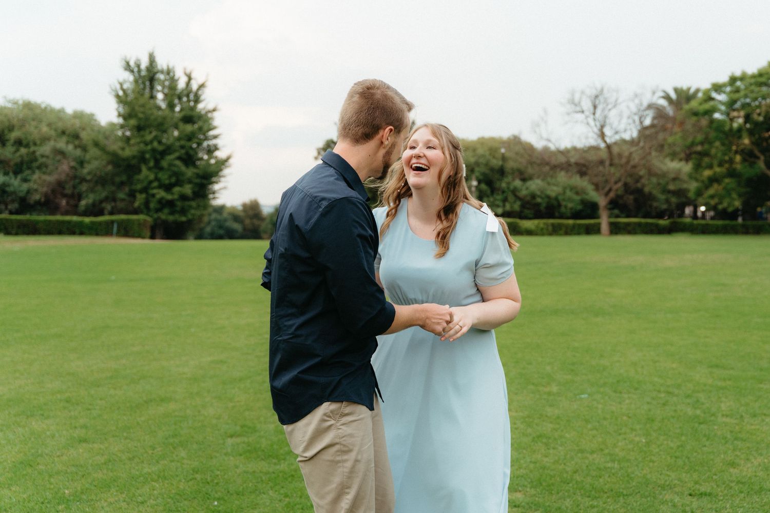 A joyful couple shares a romantic moment while laughing together on a green lawn surrounded by trees.
