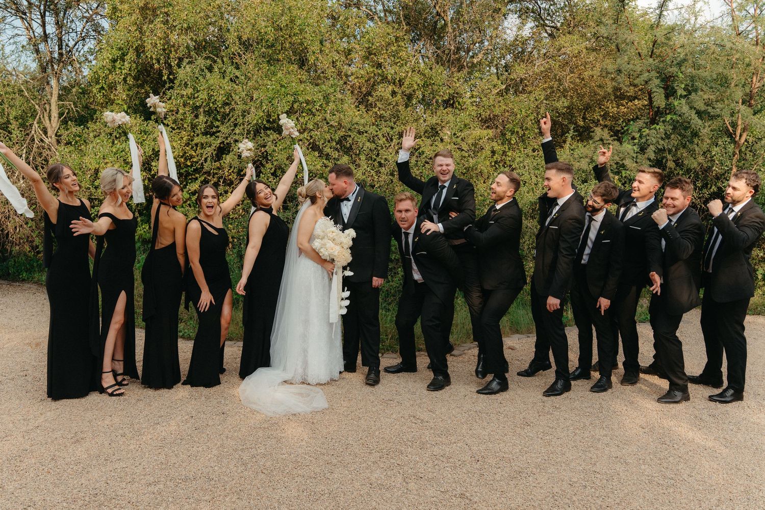 Wedding party members lined up outdoors in formal attire raise their arms in celebration against a natural backdrop.