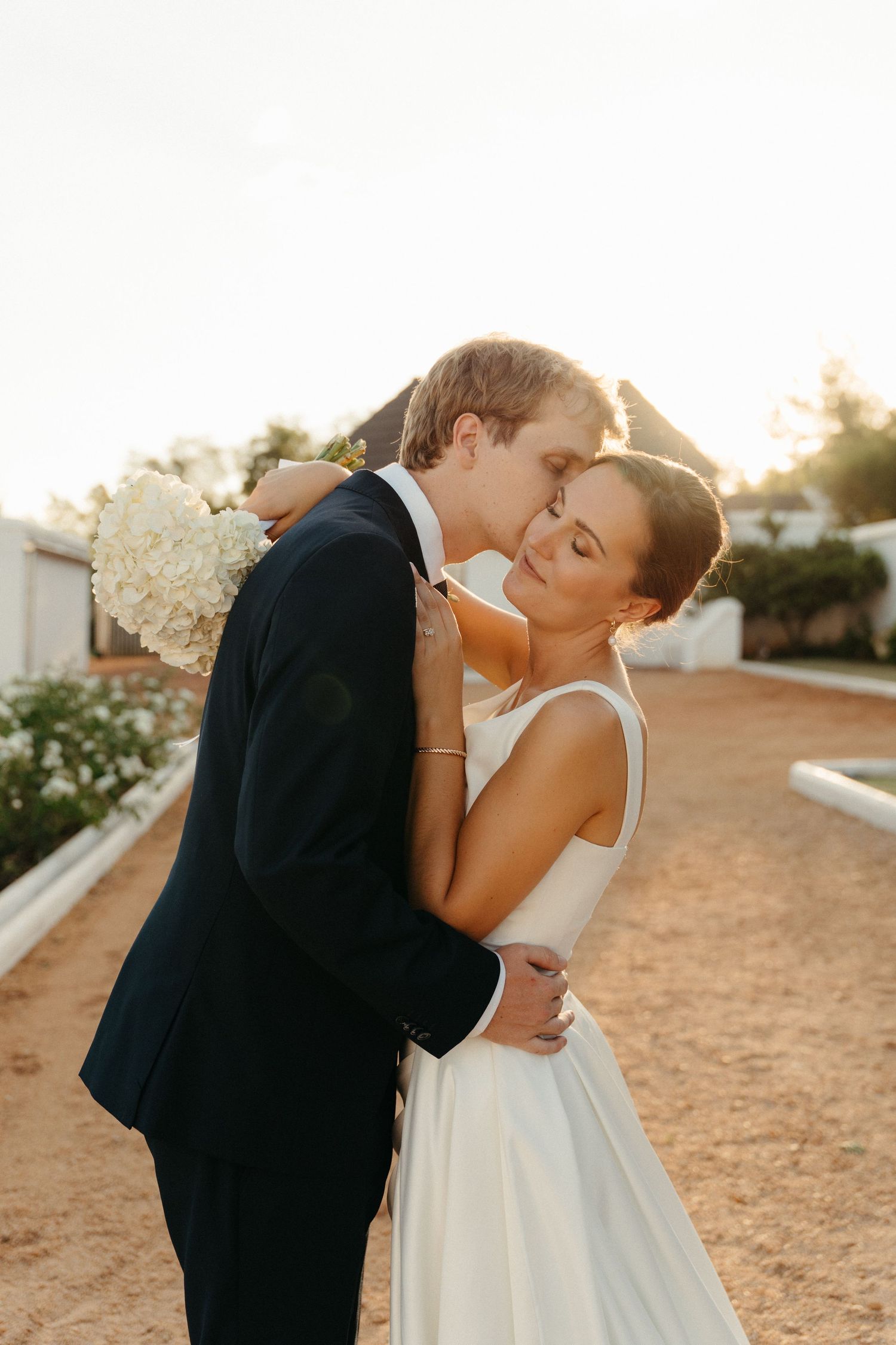 Groom tenderly embraces bride holding white hydrangeas in warm golden sunset light at Steynshoop Valley Lodge in Hekpoort, Gauteng