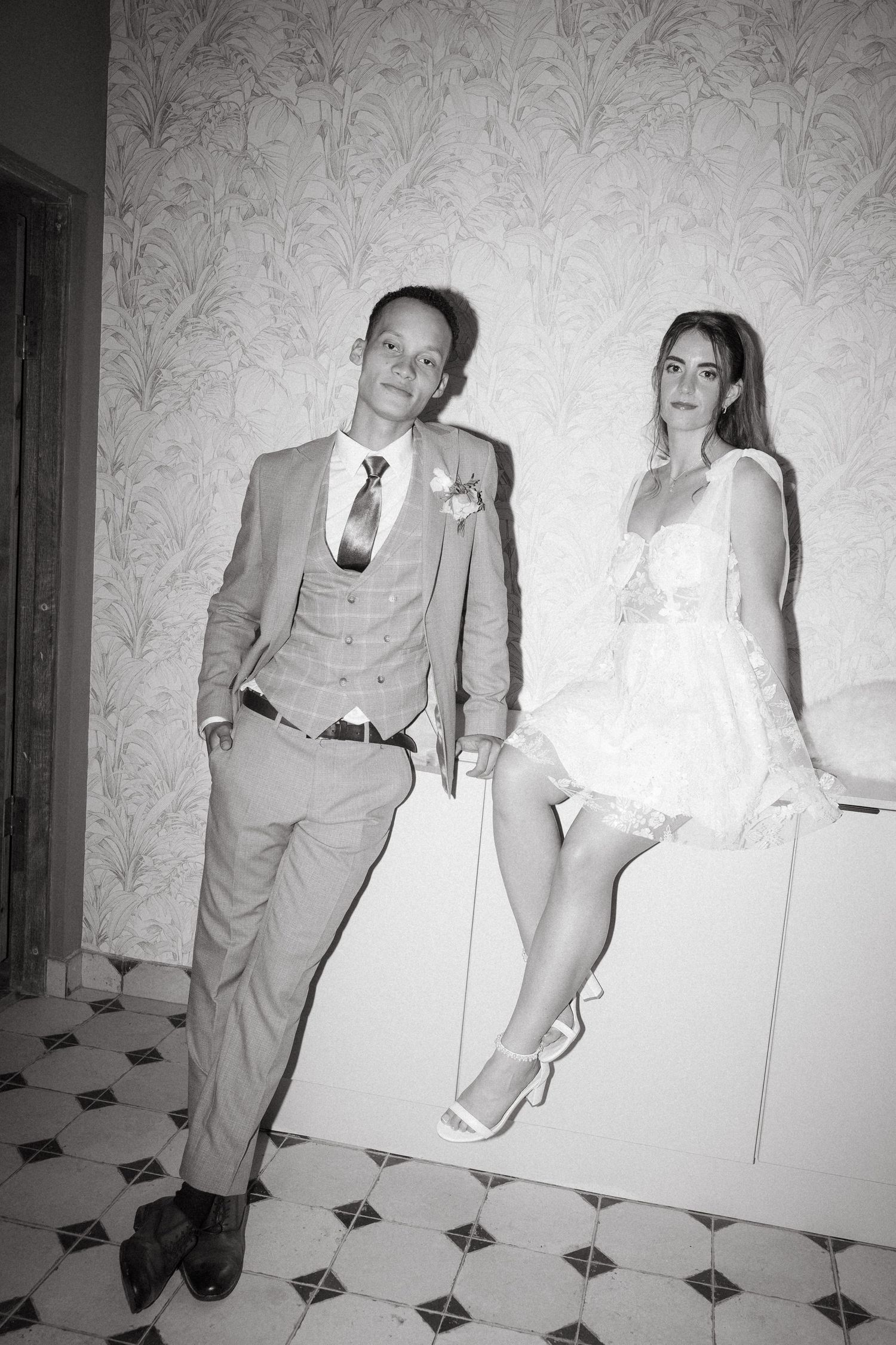Black and white flash photo of a wedding couple in formal attire, man in suit and woman in white dress, posing indoors.