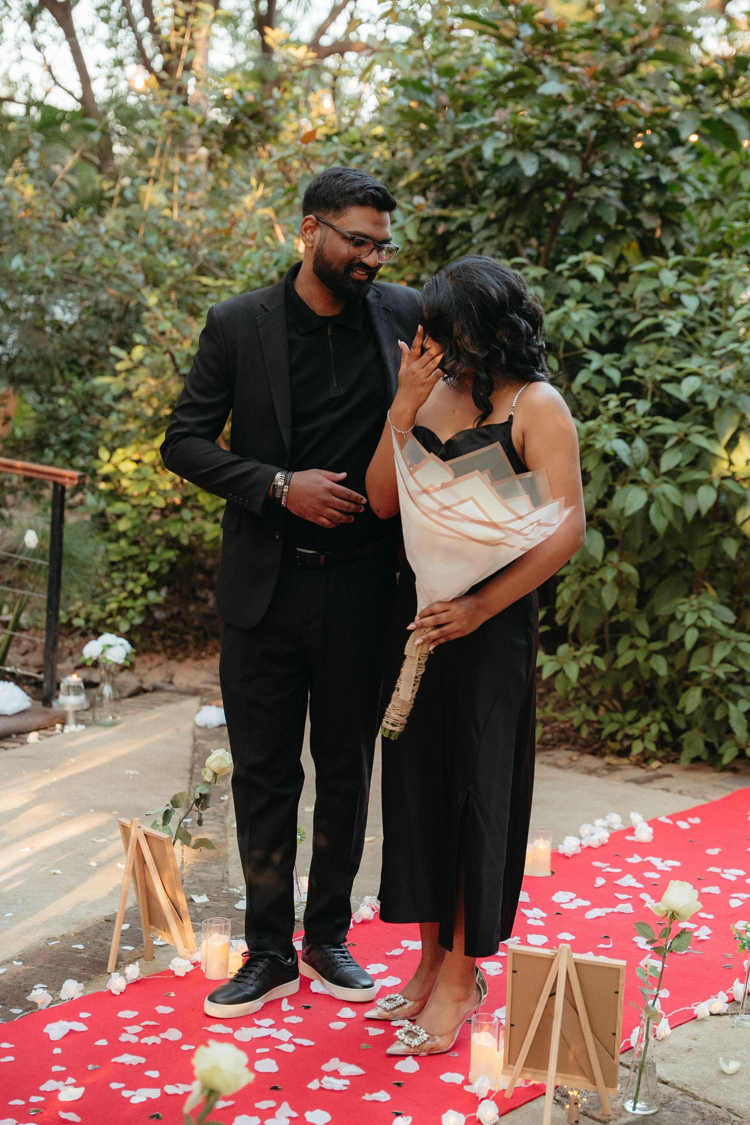 Couple sharing emotional proposal moment on red carpet with rose petals, candles, and lush greenery backdrop.