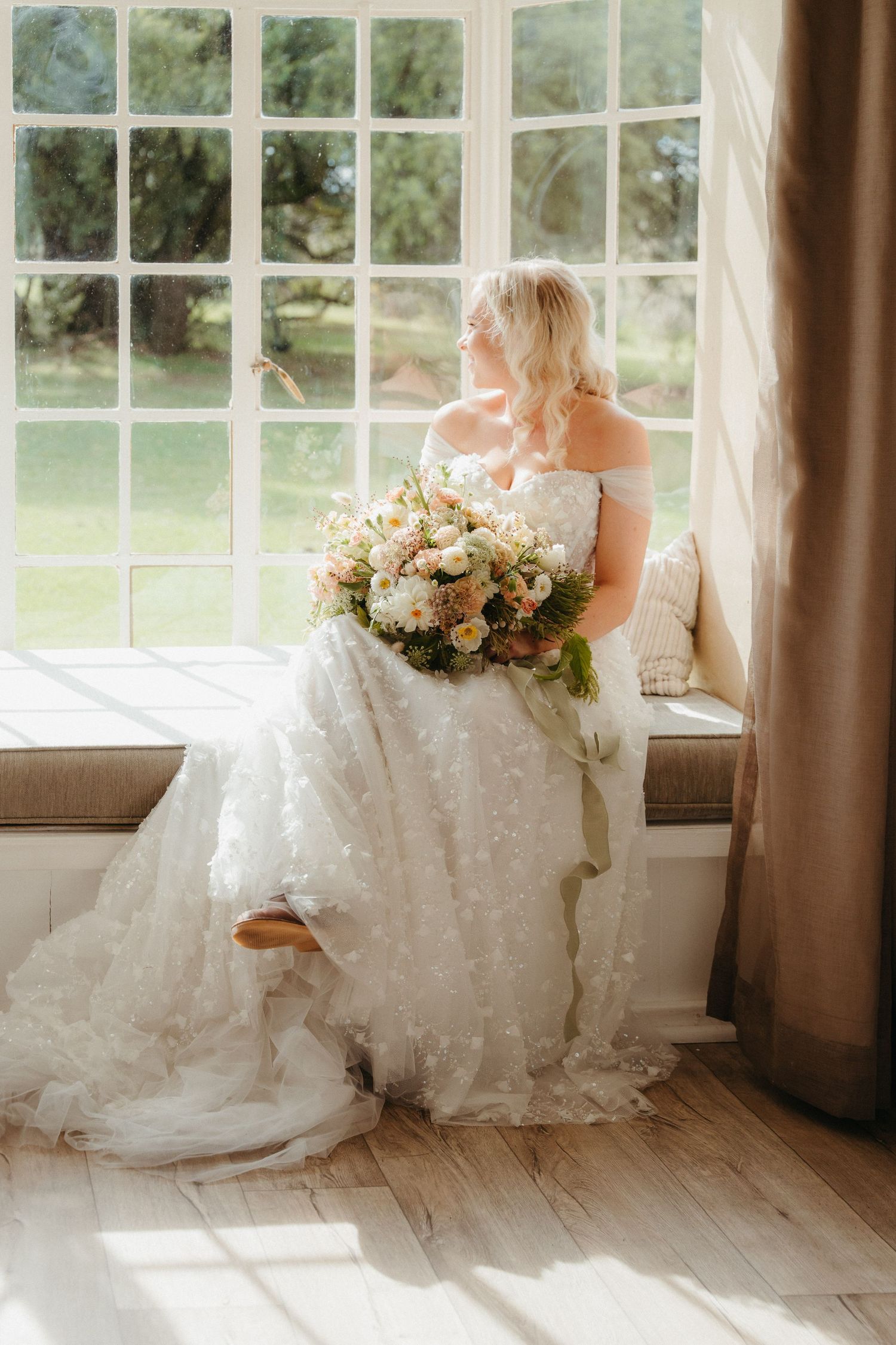 Bride in white floral gown holding bouquet sits by large window in soft natural light.