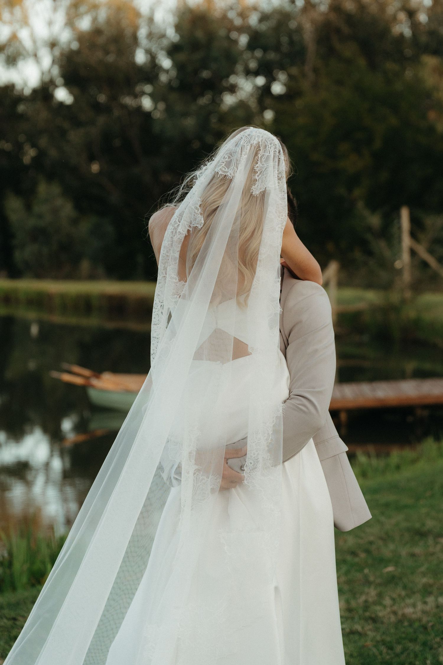 Bride and groom embracing outdoors near a lake, bride wearing a lace-trimmed cathedral veil and backless wedding gown.