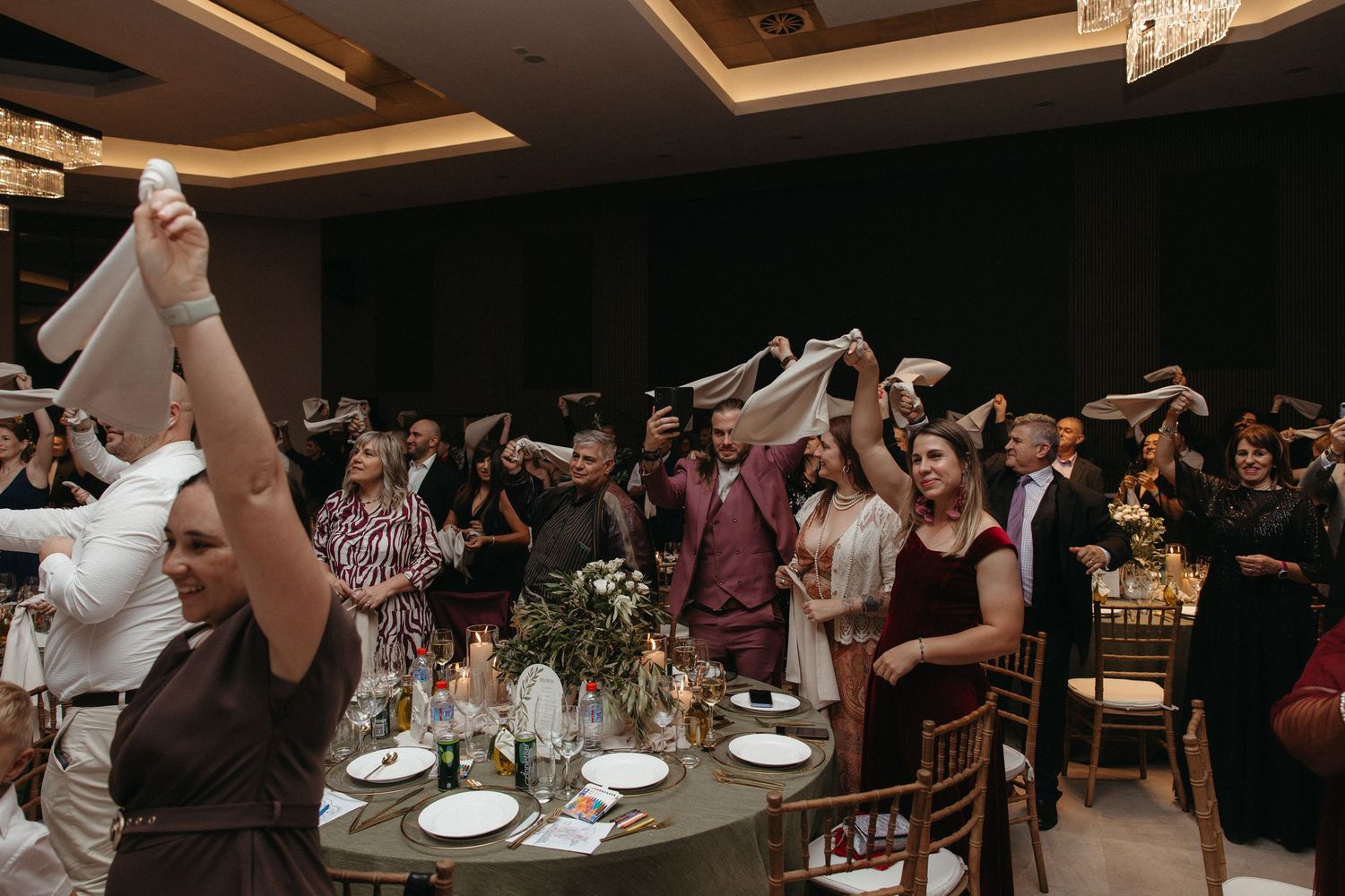 Wedding guests wave white napkins in celebration at a elegant indoor reception with candlelit tables.