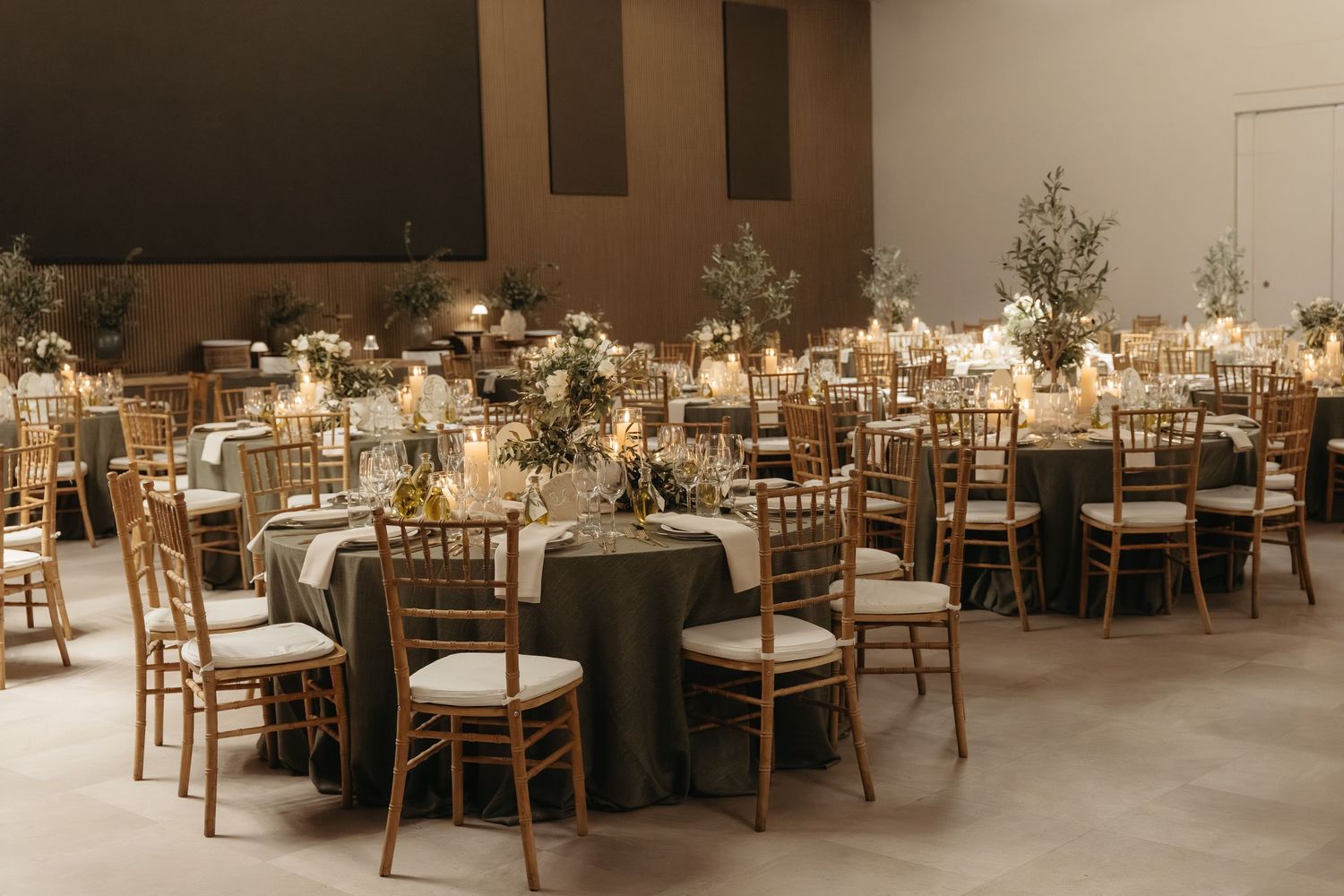 Elegant wedding reception hall with round tables, gold Chiavari chairs, dark linens, and floral centerpieces in warm candlelight.