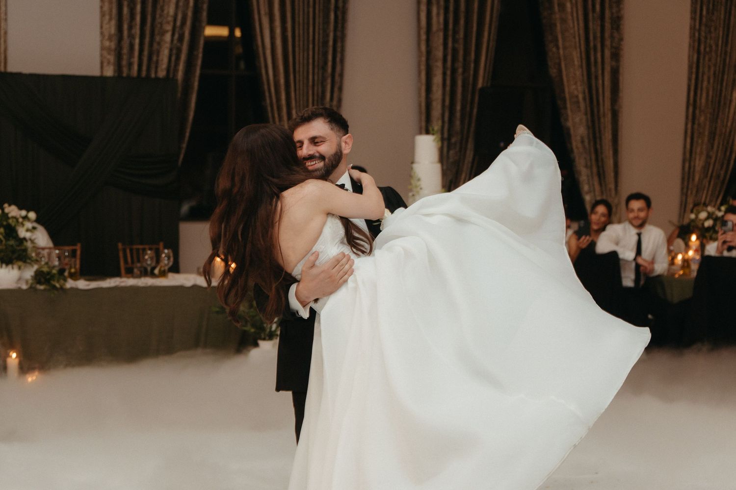 Groom lifting bride in white gown during romantic first dance at elegant wedding reception.