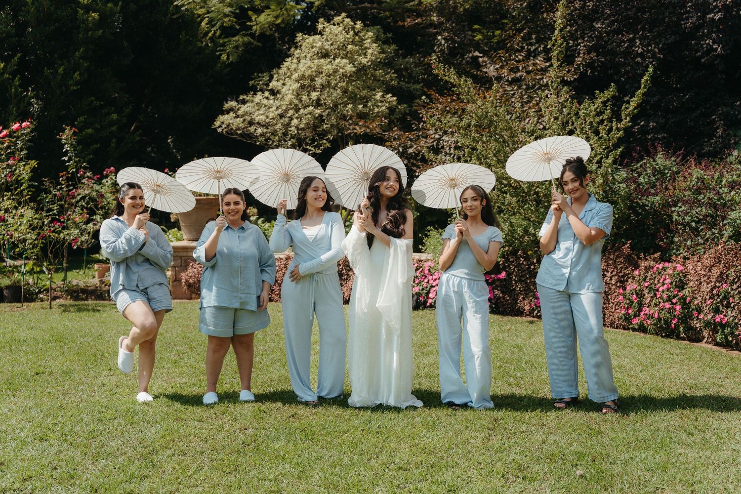 Bride in white ao dai with bridesmaids in light blue suits holding parasols in a lush garden wedding photo.