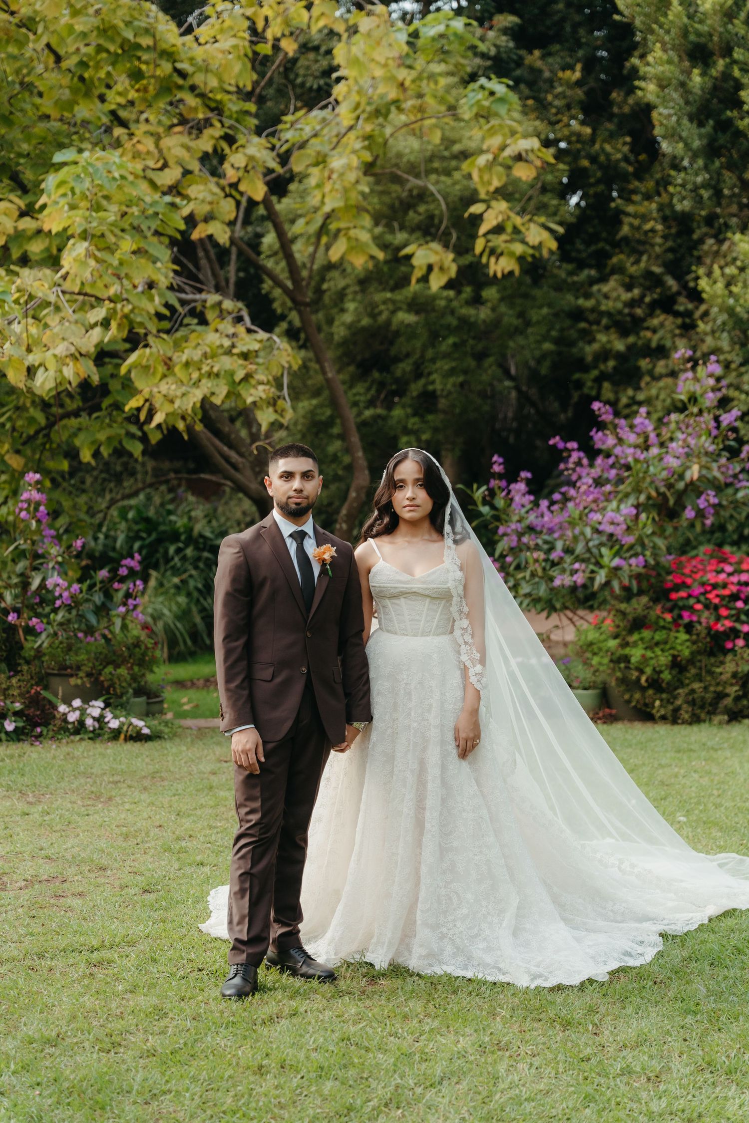Bride in beaded gown with veil and groom in brown suit pose together in lush garden with colorful flowers.