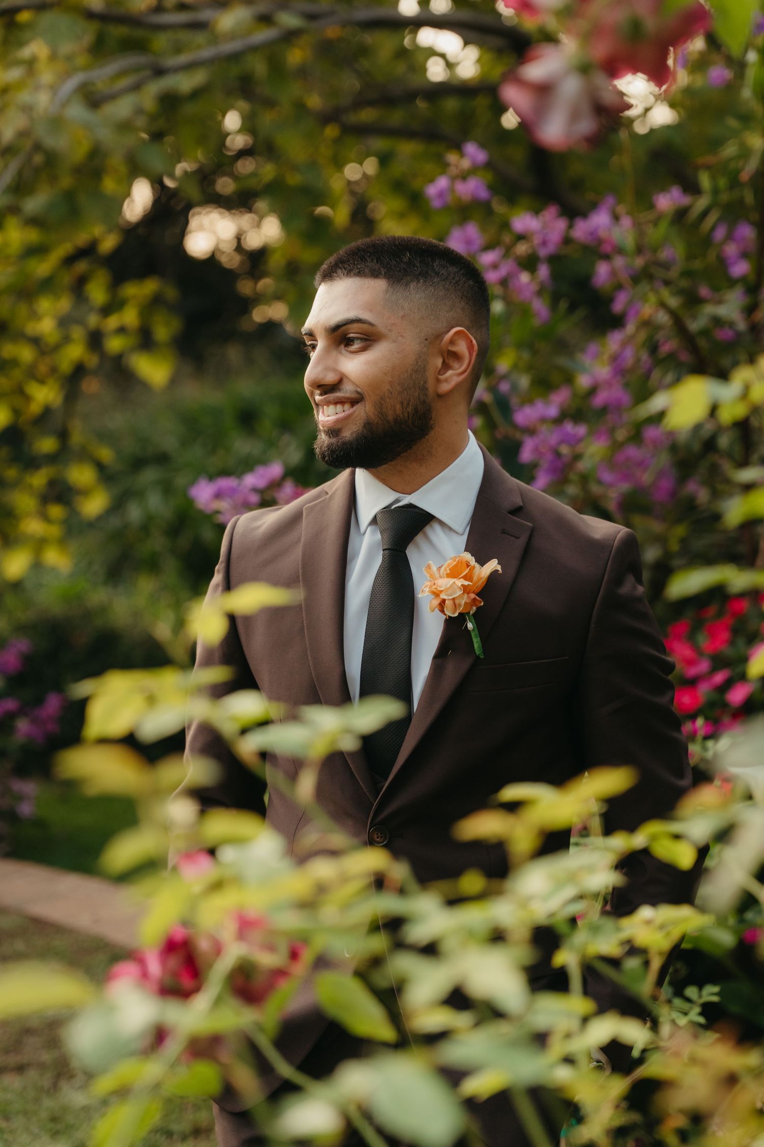 Smiling groom in brown suit with orange boutonniere standing among colorful garden flowers.
