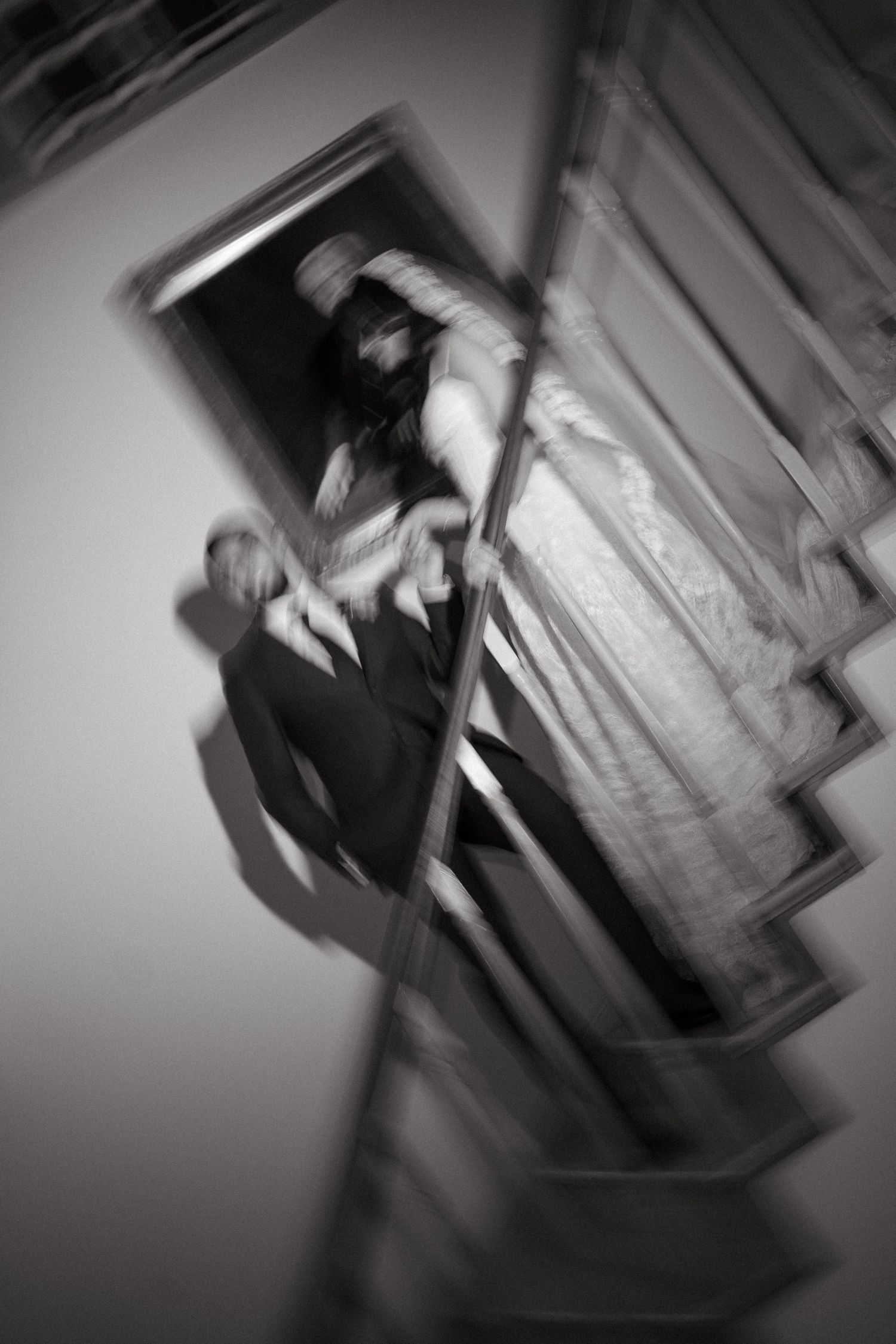 Blurred black and white photo of elegantly dressed figures near ornate frames, creating a surreal, motion-blur artistic effect.