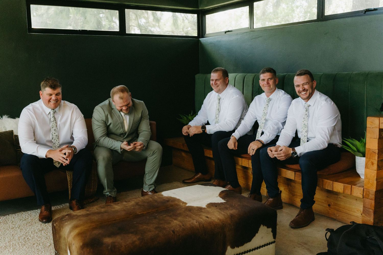 Five groomsmen in formal attire sit together on leather seating in a dark room during wedding preparations.