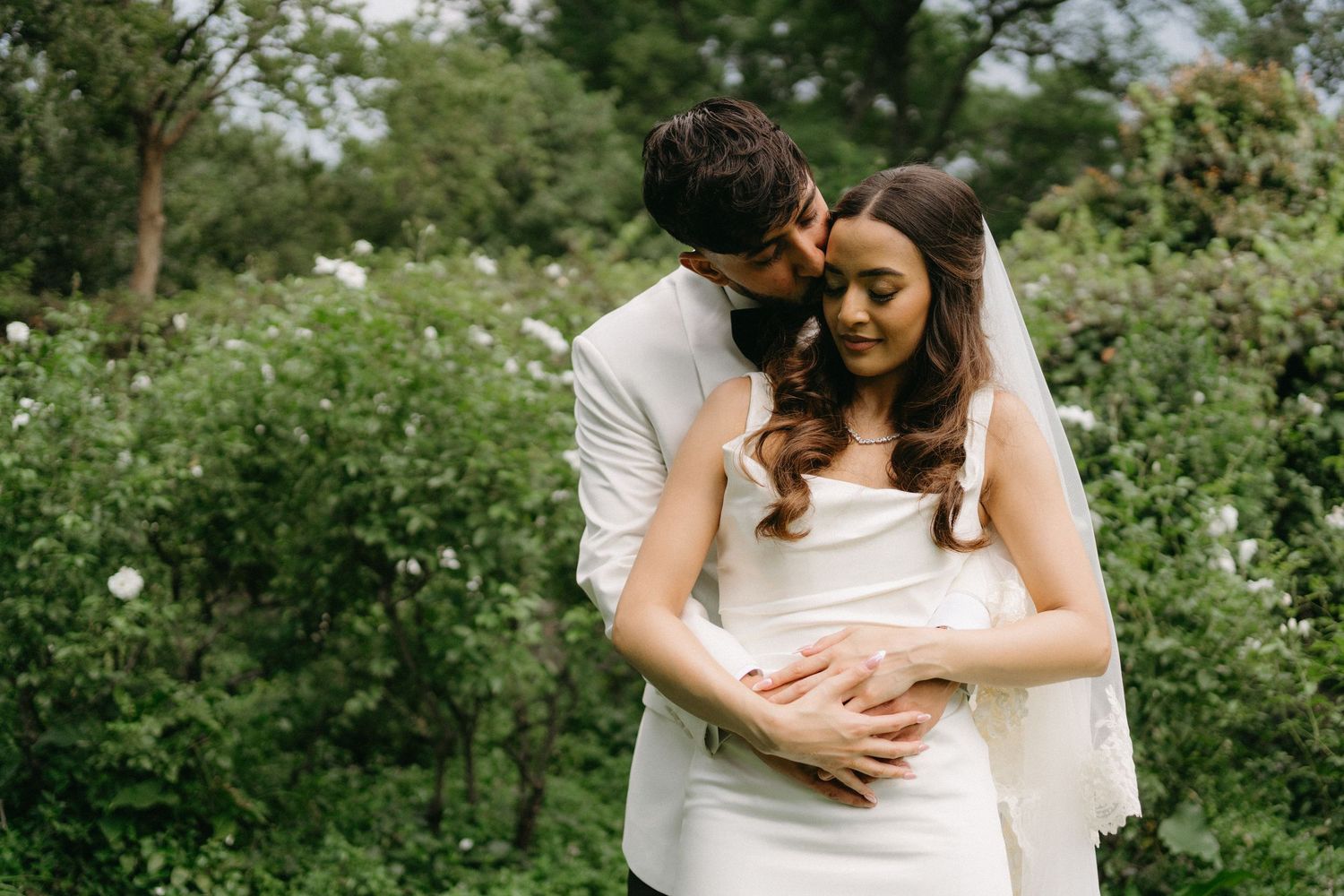 Romantic garden wedding portrait of newlyweds embracing among lush greenery.