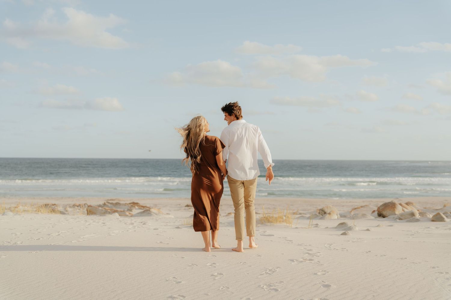 Couple walking hand in hand on a sunny beach toward the ocean.