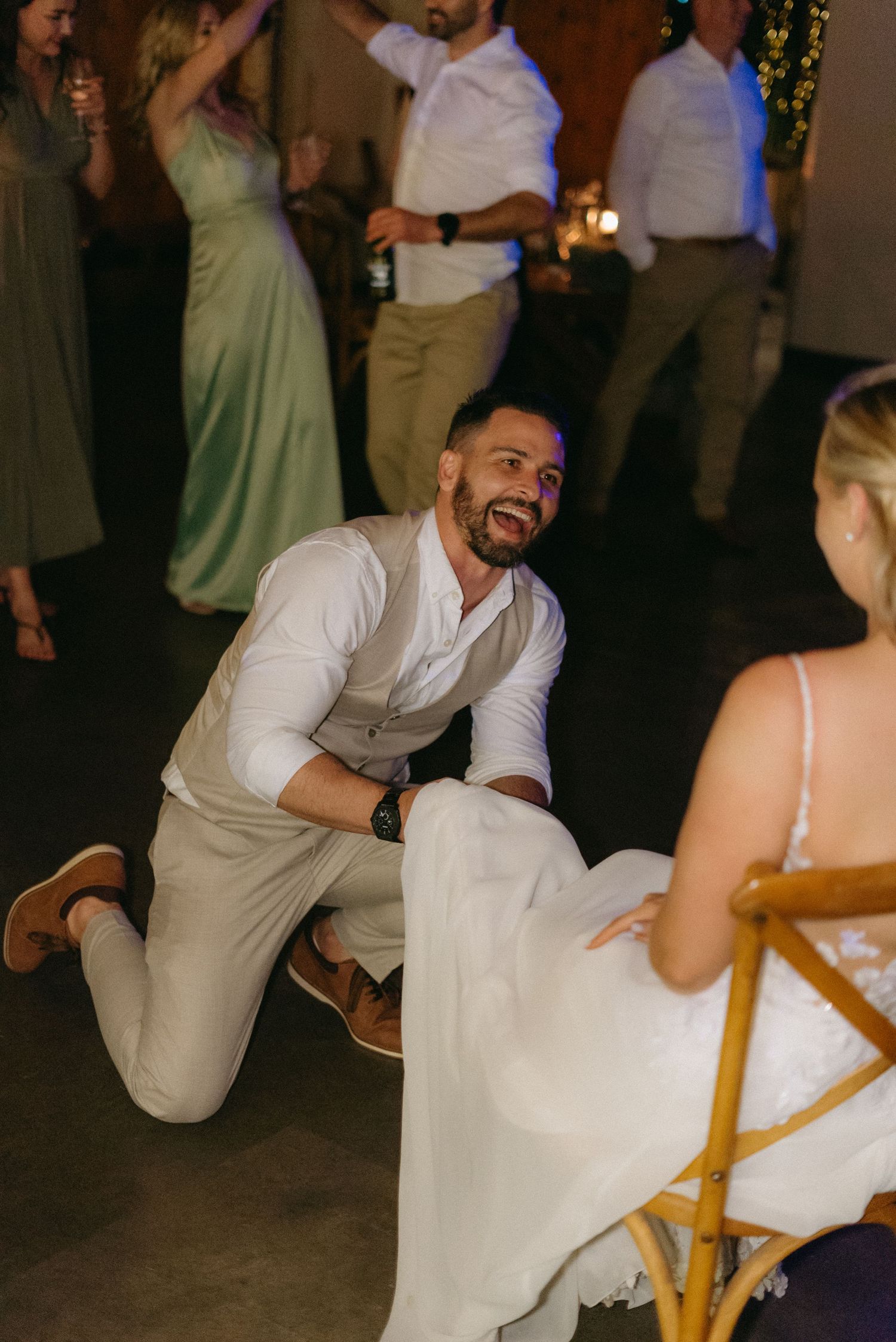 Groom kneels playfully in front of seated bride during wedding reception dance floor game.