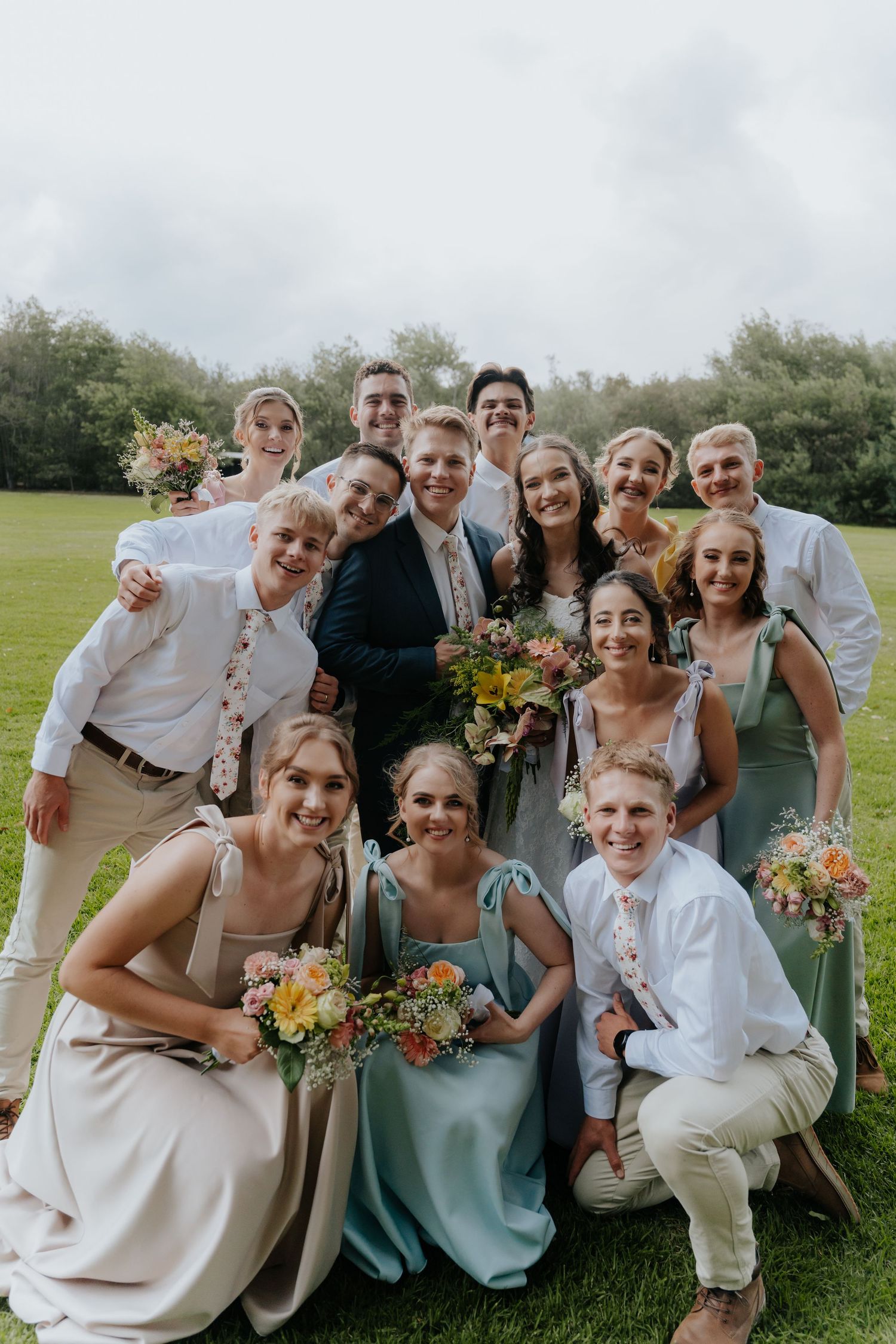 Wedding party poses together on a grassy lawn with colorful bouquets and mix of sage green and neutral colored attire.
