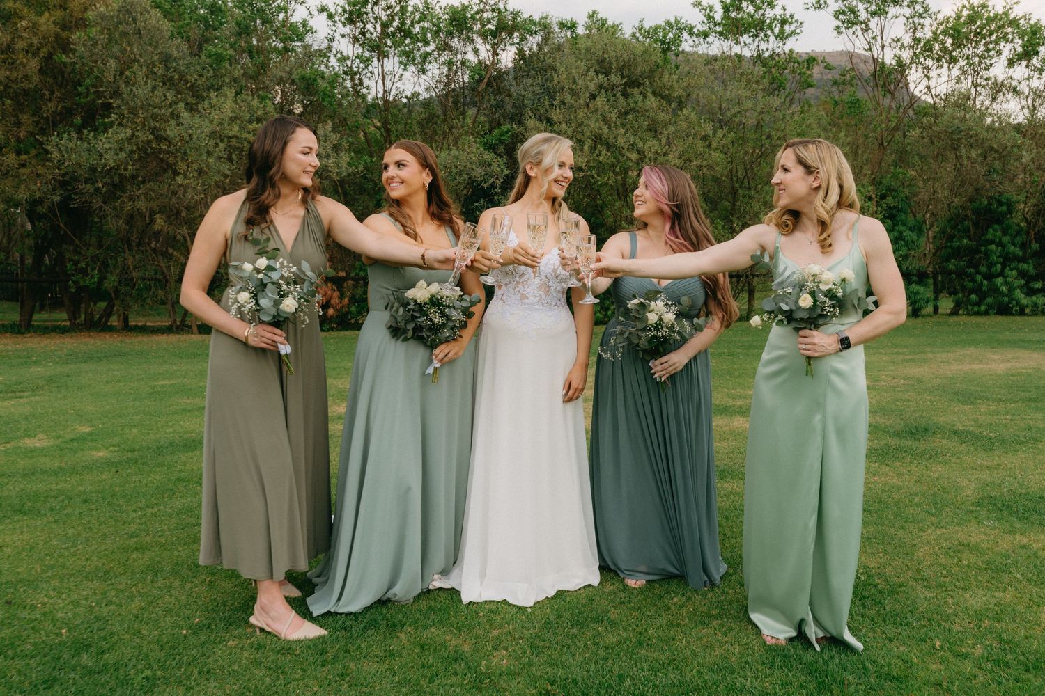 Wedding party in sage green bridesmaid dresses poses together on a grassy lawn.