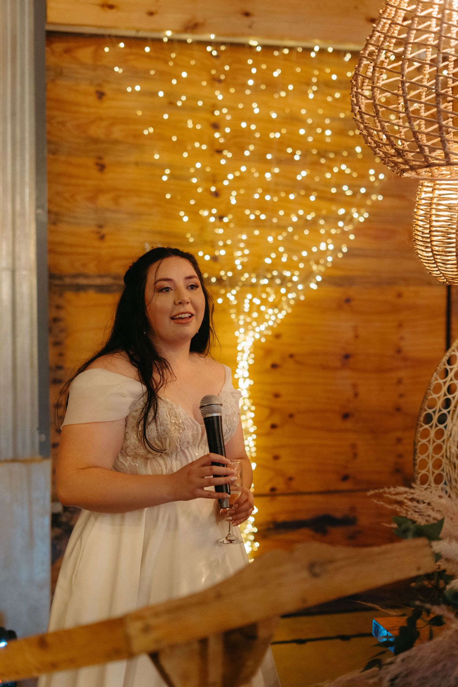 A person holds a microphone while giving a speech in a rustic wood venue decorated with twinkling string lights.