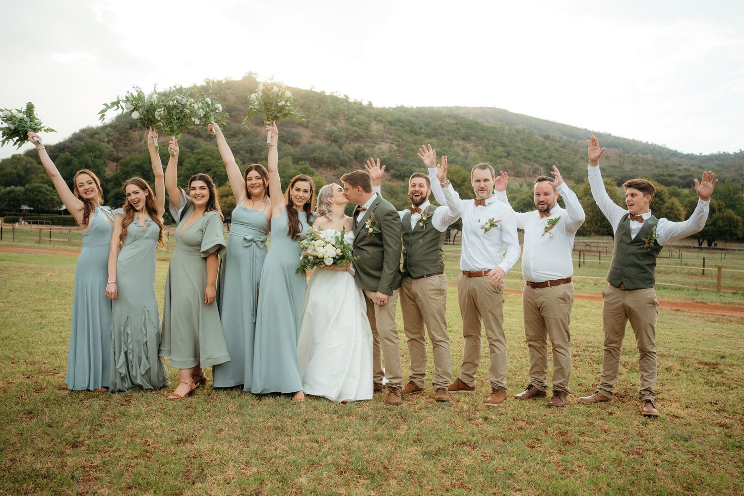 Wedding party celebrates with raised arms in a rustic outdoor setting with rolling hills in the background.