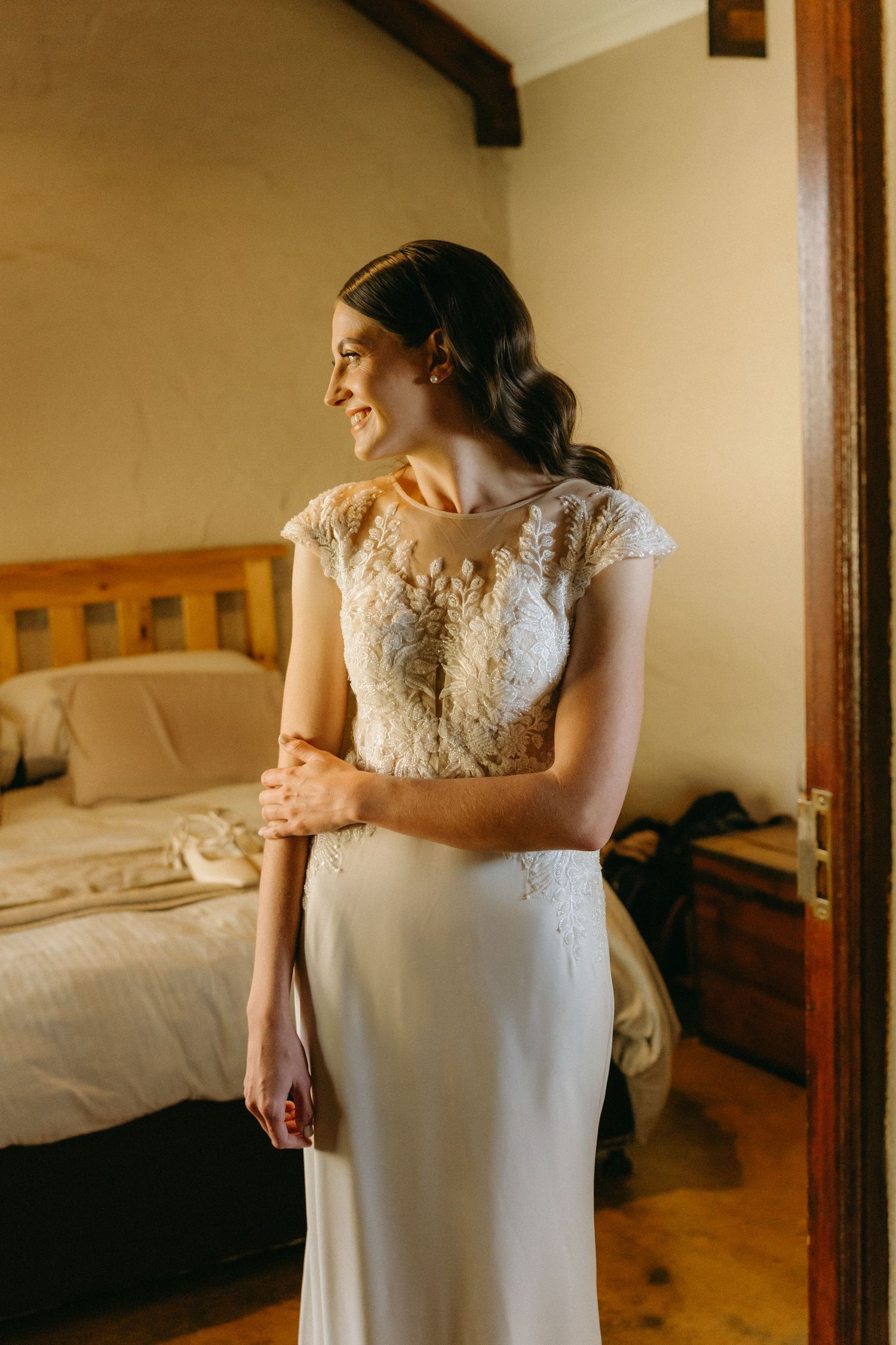 Elegant white wedding dress with lace details shown in a rustic bedroom setting.