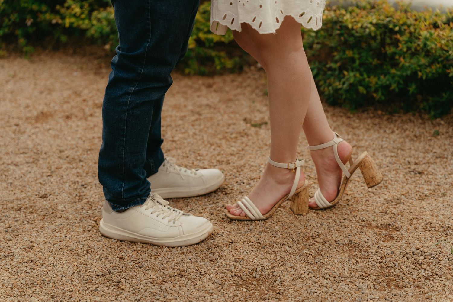 Close-up view of feet and shoes on gravel path showing white sneakers and nude heels.