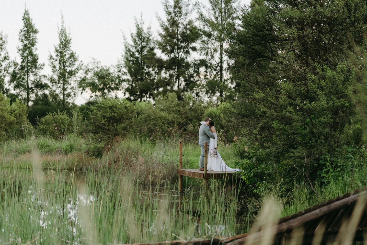 A couple stands on a wooden deck surrounded by tall pine trees and natural grasses beside a lake.