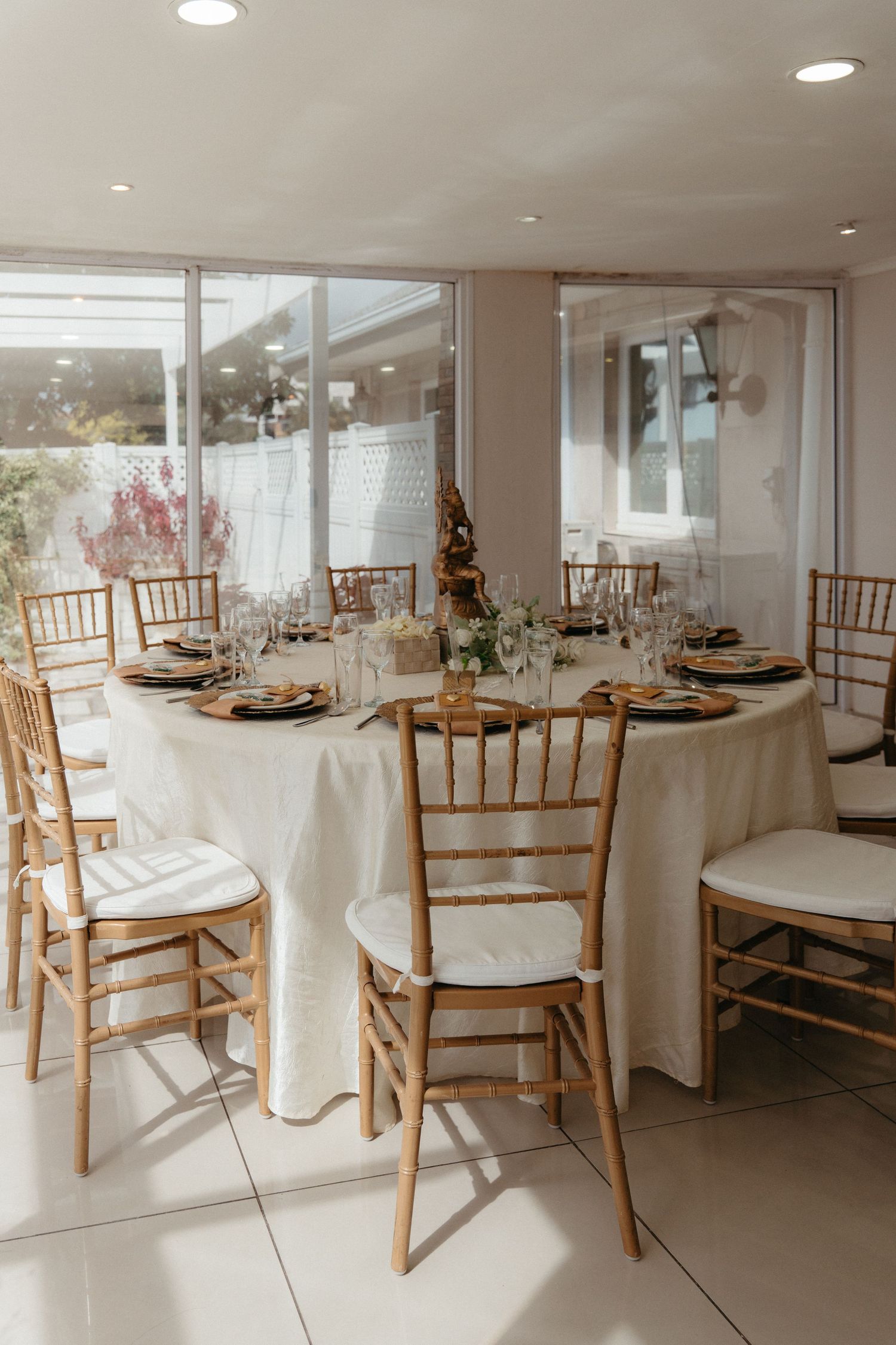 Elegant wedding reception venue with white tablecloths and wooden chiavari chairs in a bright, sunlit room.