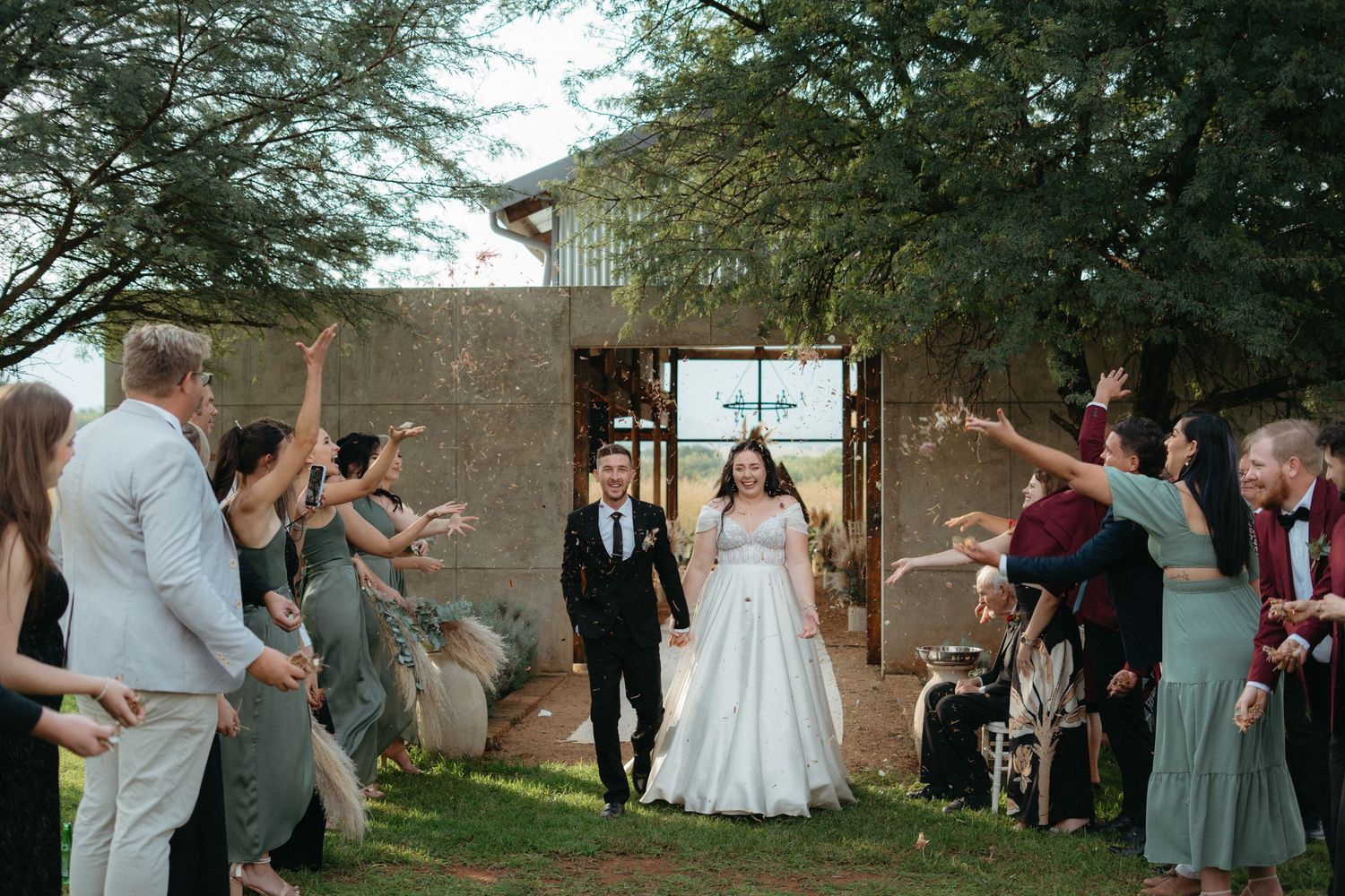 Wedding couple exits through a rustic stone archway as guests celebrate with raised arms at an outdoor ceremony.