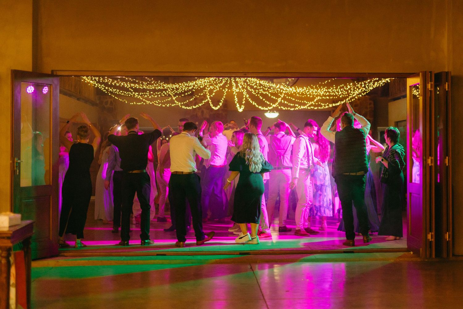 Wedding venue decorated with string lights and filled with dancing guests in formal attire.