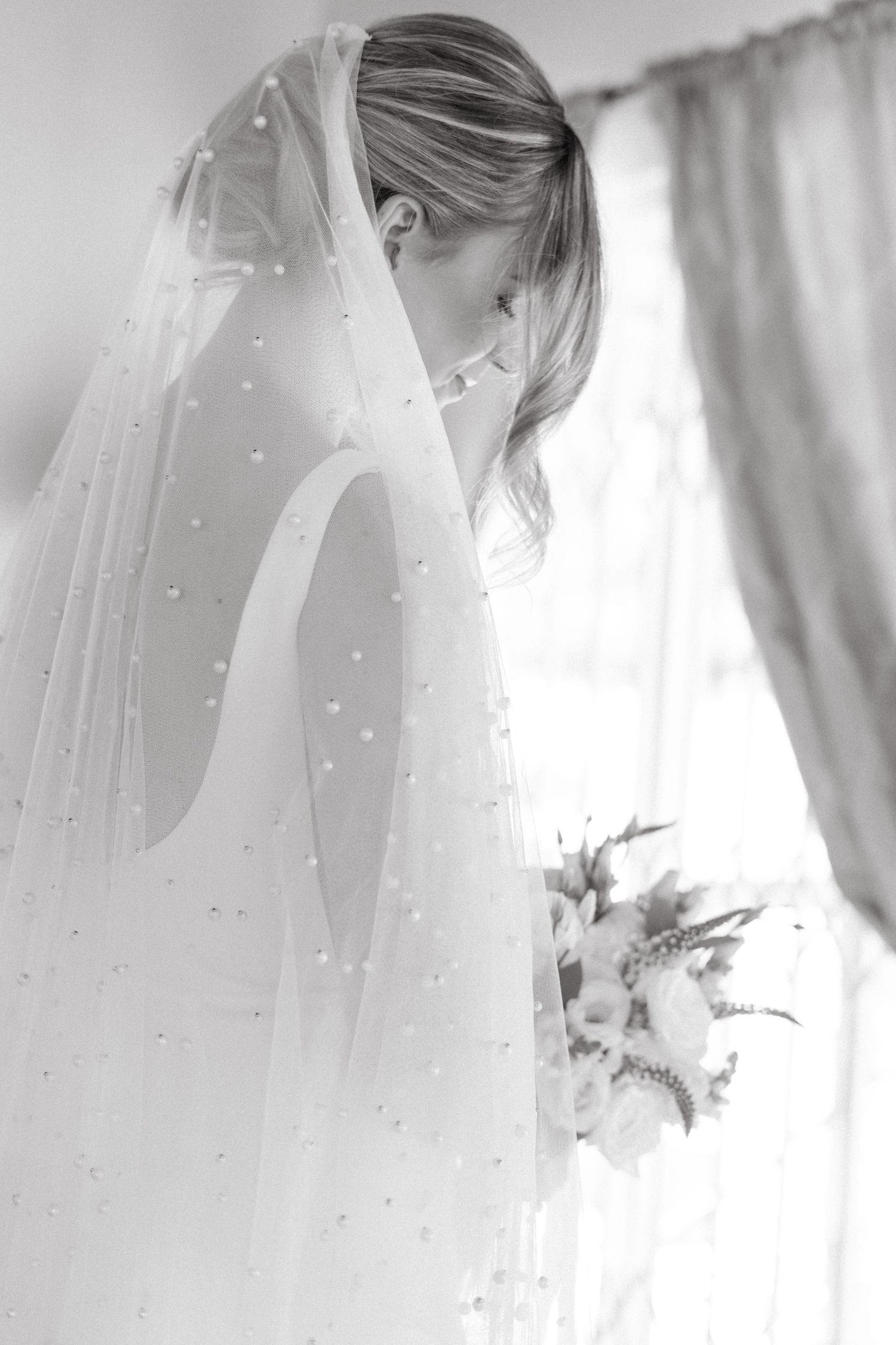 Black and white photo of bride in delicate pearl-studded veil holding bouquet near window in soft lighting.