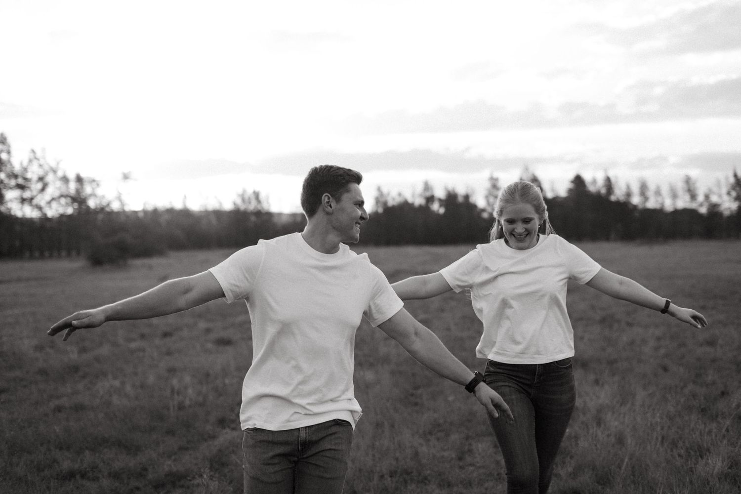 Two people in white shirts run with outstretched arms through a grassy field at dusk in black and white.