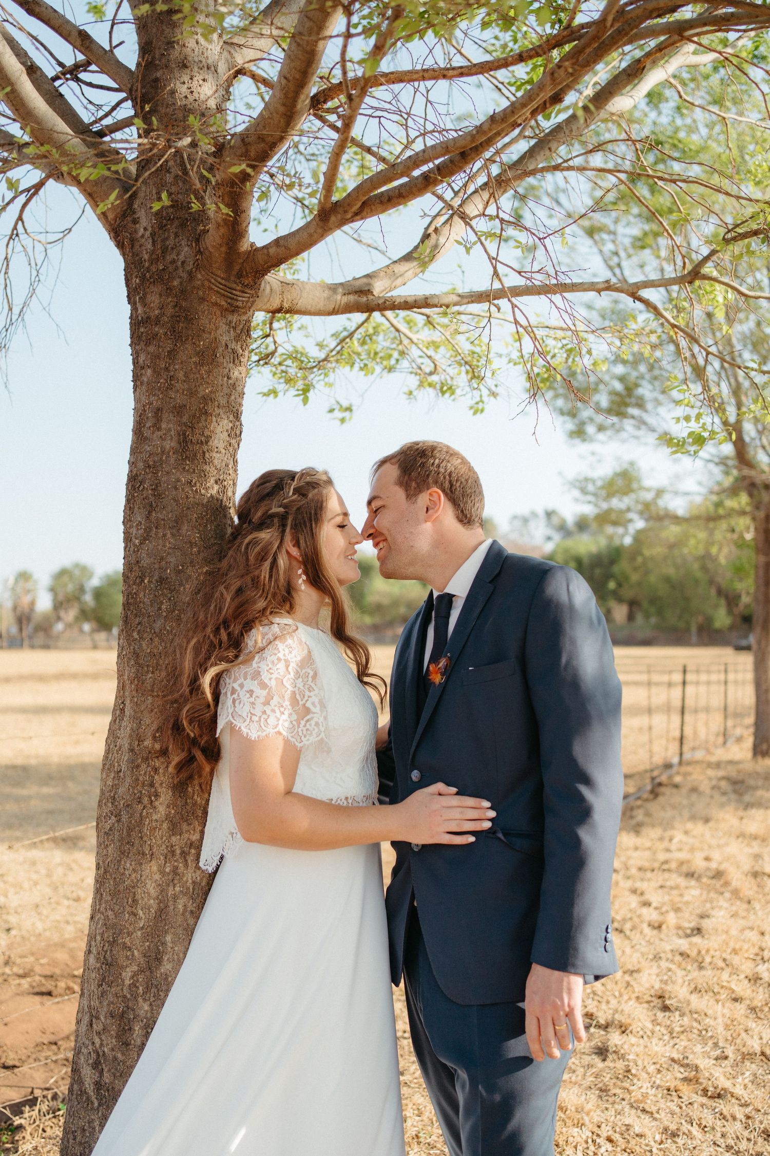 A tender wedding moment captured under a large tree in a rustic outdoor setting.