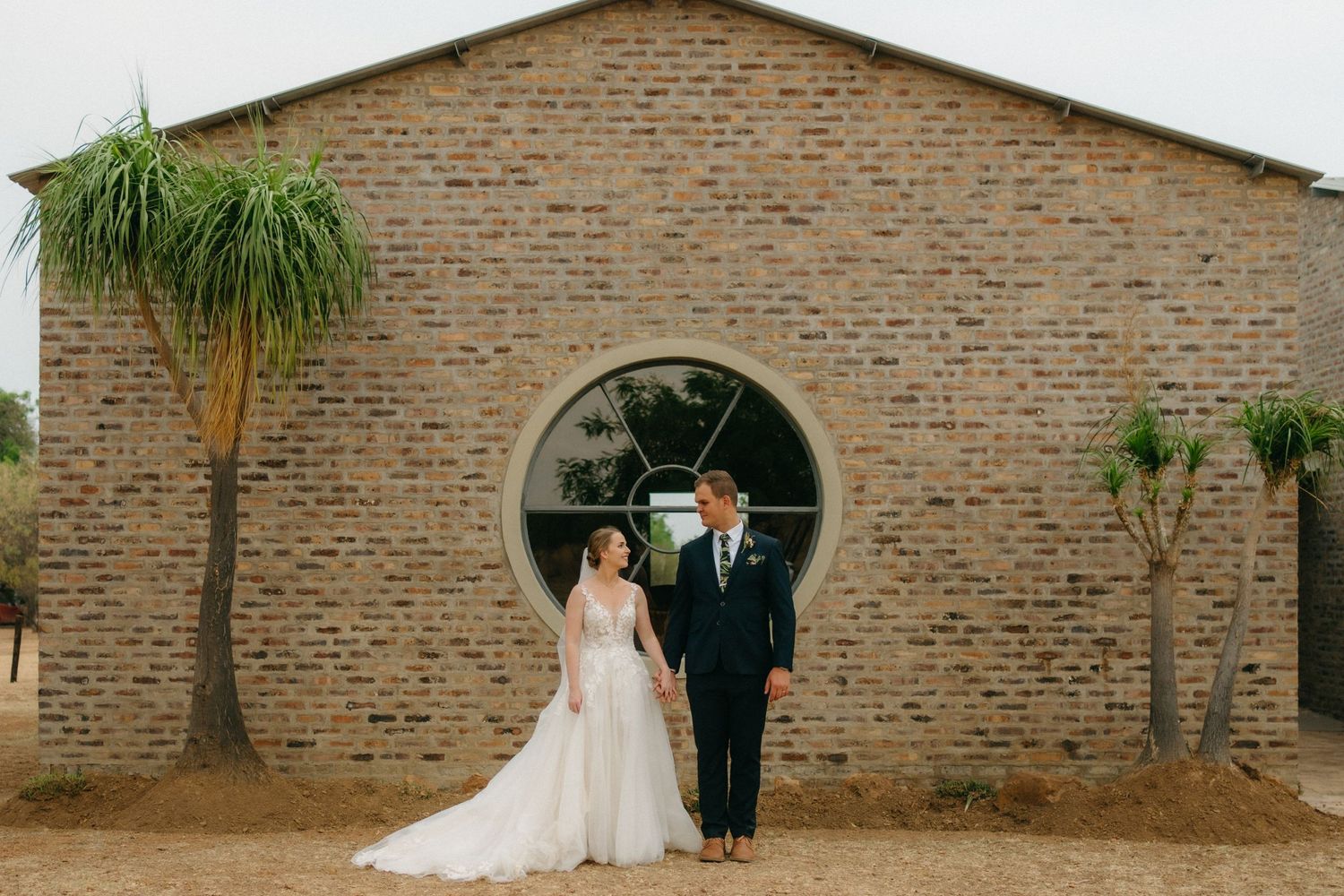 A wedding couple poses in front of a brick building with a circular window and palm trees.
