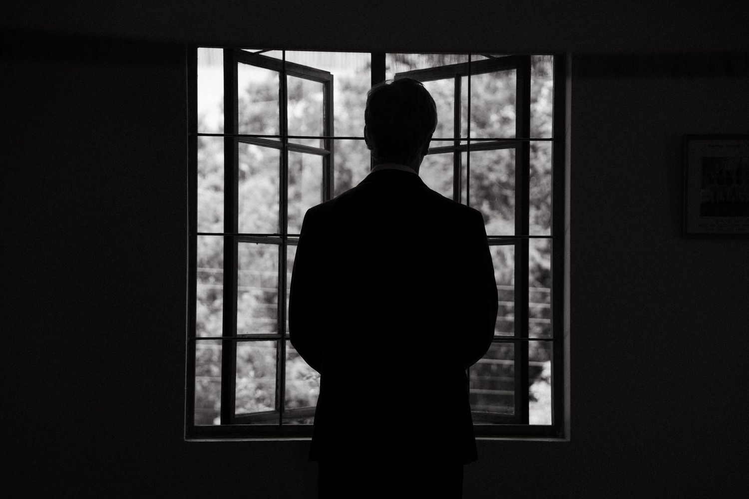 A silhouetted figure stands in darkness near a window, creating a moody black and white composition.