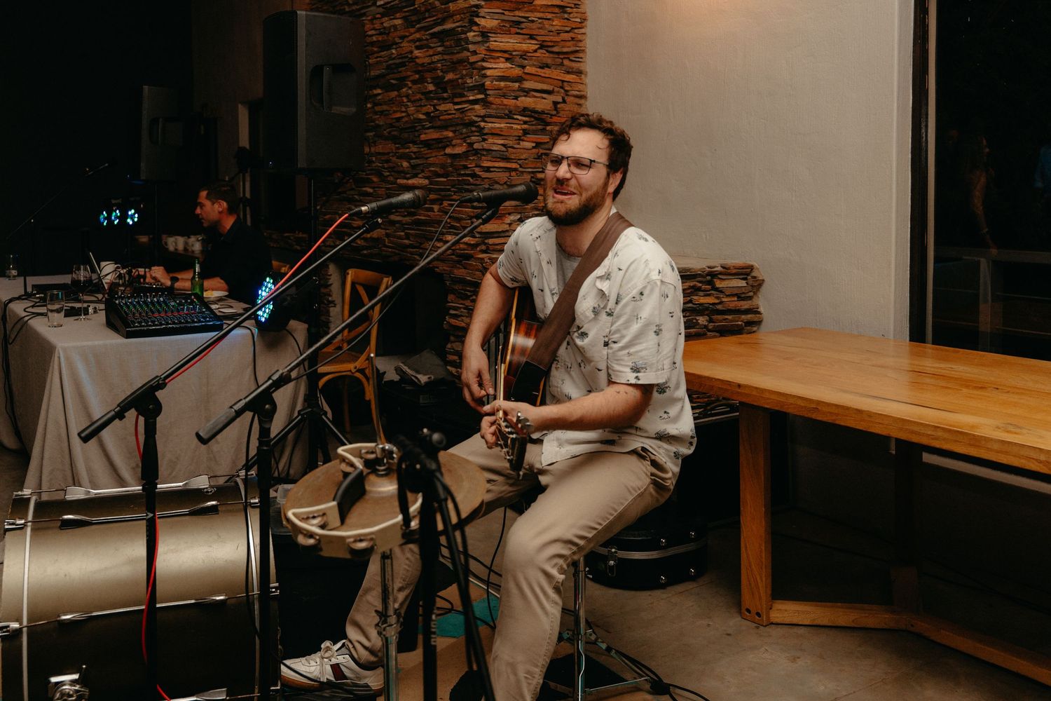 A musician performs with an acoustic guitar while seated in a modern venue with stone wall accents.