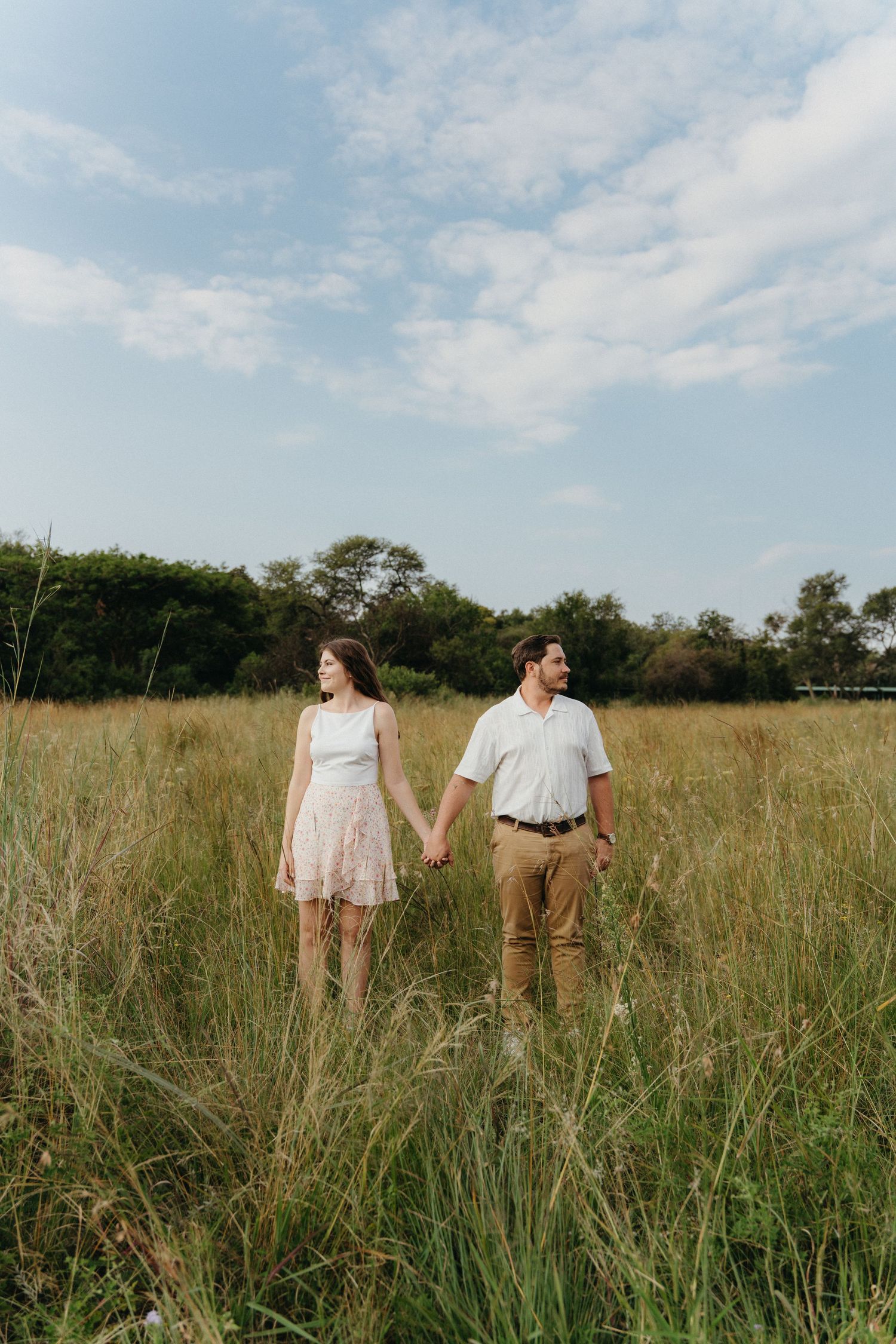 Couple holding hands in tall grass field wearing casual summer outfits with trees and blue sky behind them.