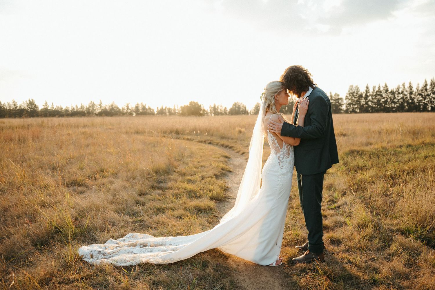 Bride and groom embracing in golden field at sunset, bride wearing lace wedding dress with long veil.
