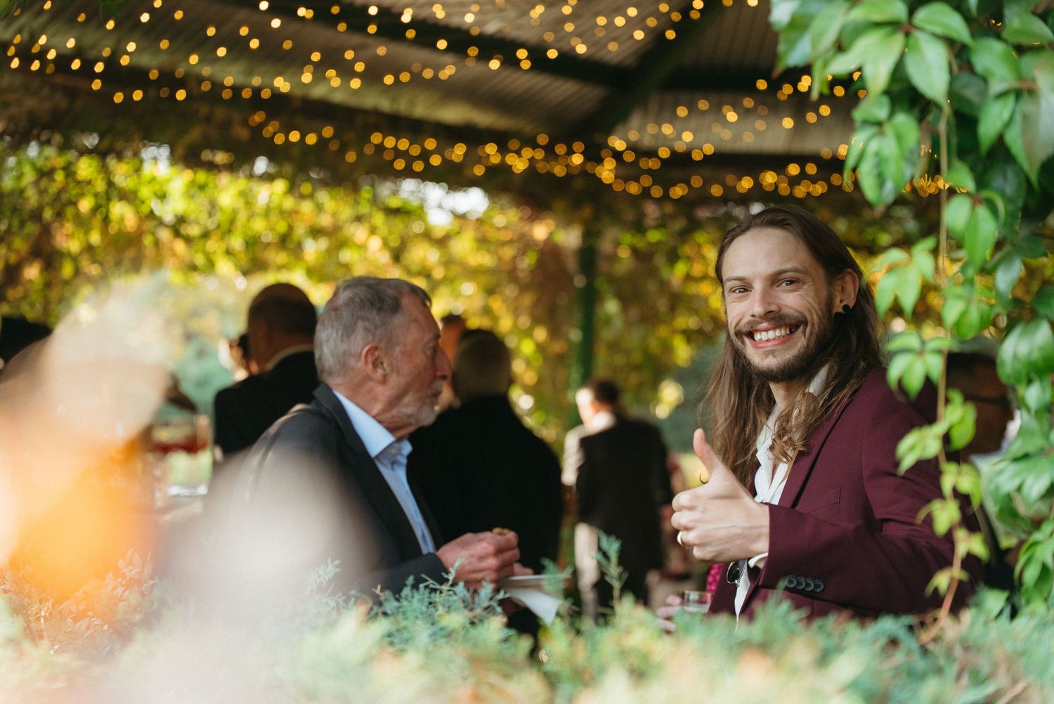 Two men smiling at an outdoor garden event with string lights and lush greenery in the background.