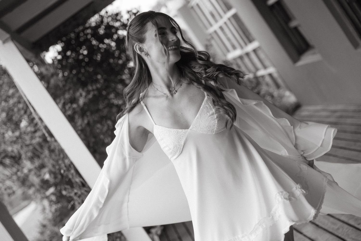 Black and white photo of a bride in a flowing white robe and open shirt, hair blowing, on a porch.